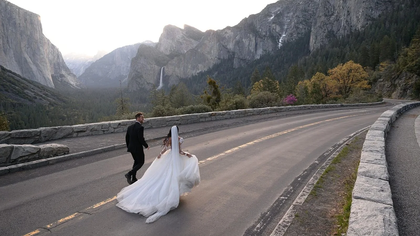 A bride in a white wedding dress and veil and a groom in a black suit walking along a mountain road with a scenic canyon, tall cliffs, trees with autumn foliage, and a waterfall in the background in Yosemite National Park