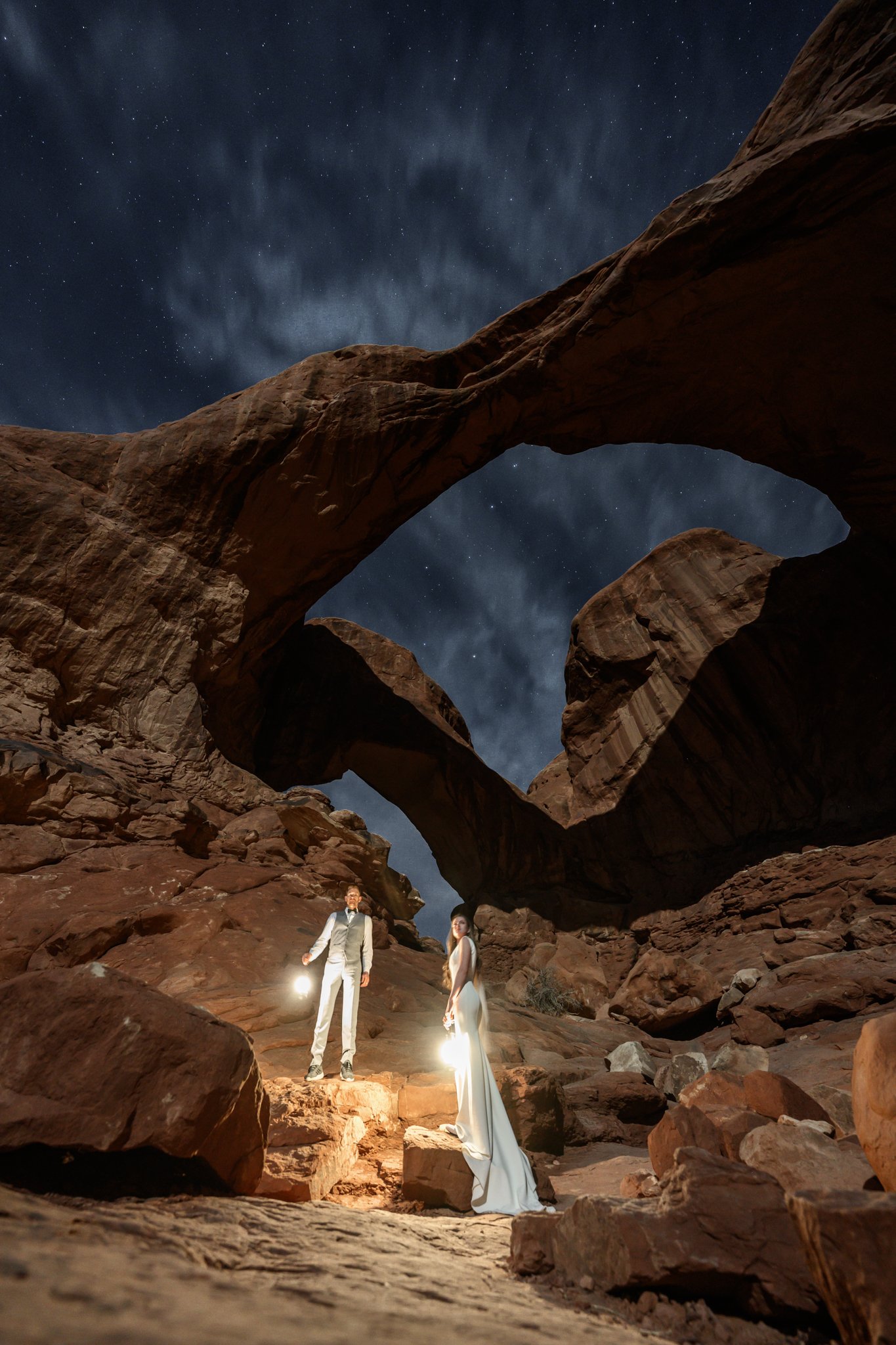 a bride and groom holding lanters at night under the double arch in Arches National Park in Moab, Utah