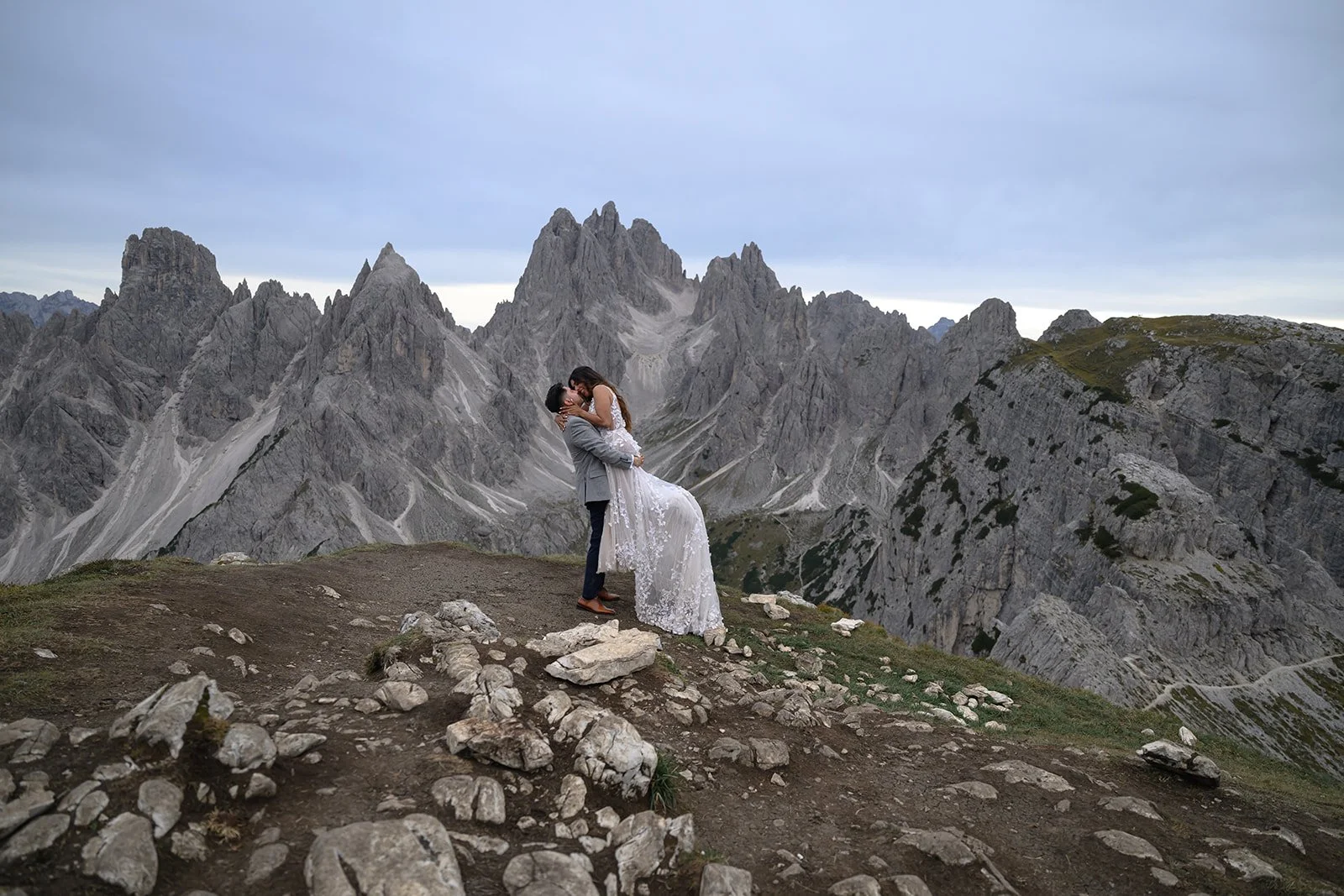 a bride and groom in the dolomites in italy with a mountain range behind them, the groom is picking up the bride and they are kissing