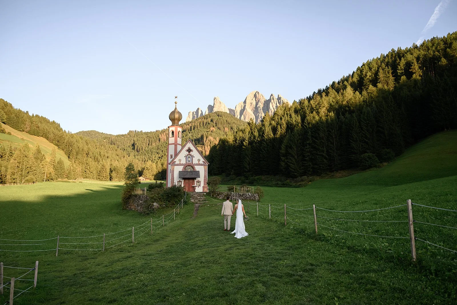 a photo of a bride and groom walking towards a small church in a valley in the dolomites with mountains peaking up behind the hill in the distance
