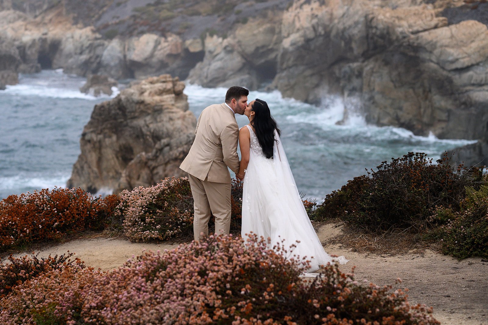 a photo of a bride and groom kissing on a cliffside in big sur with flowers around them and the ocean behind them