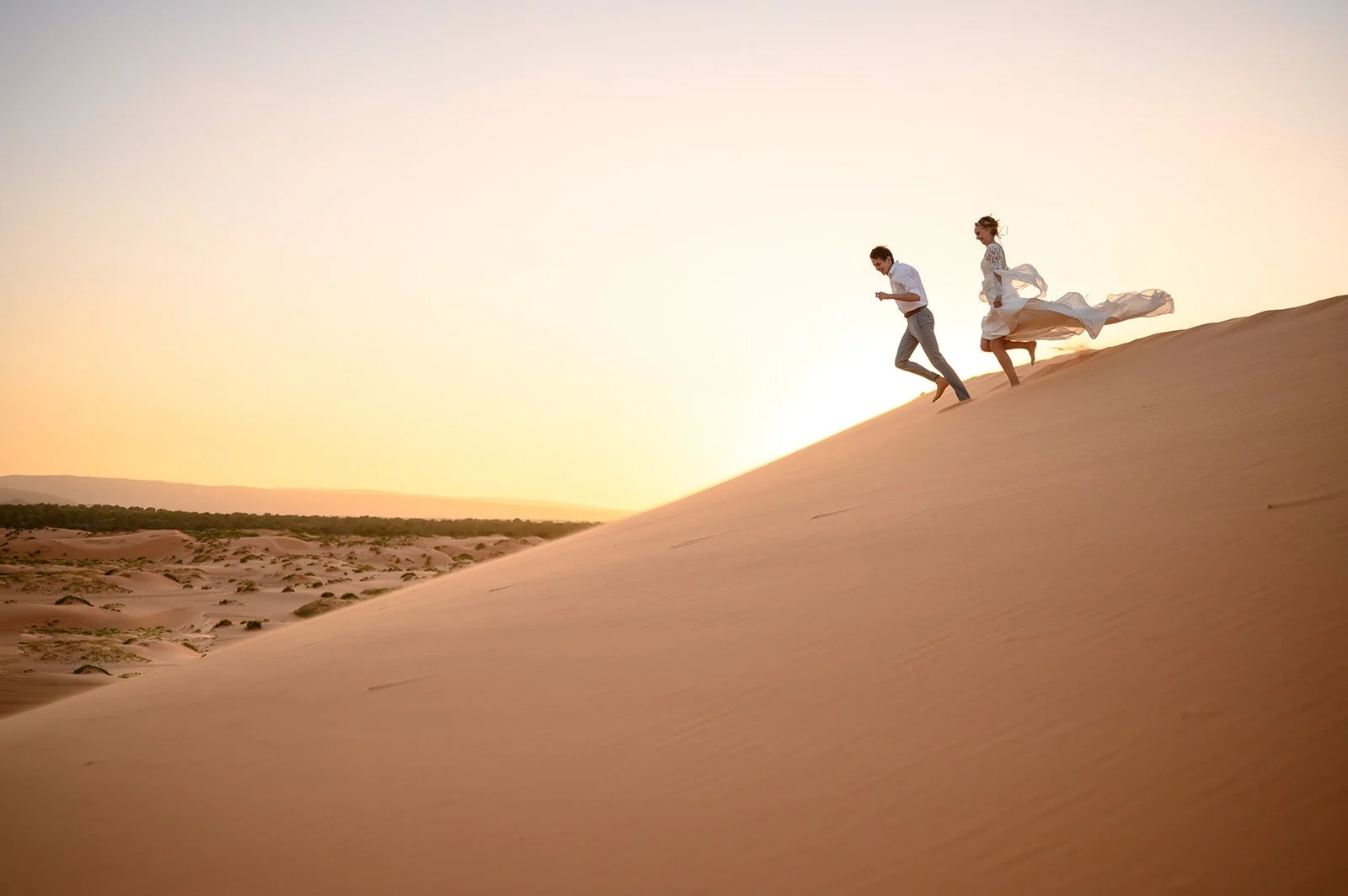 a bride and groom running down a sand dune at sunset