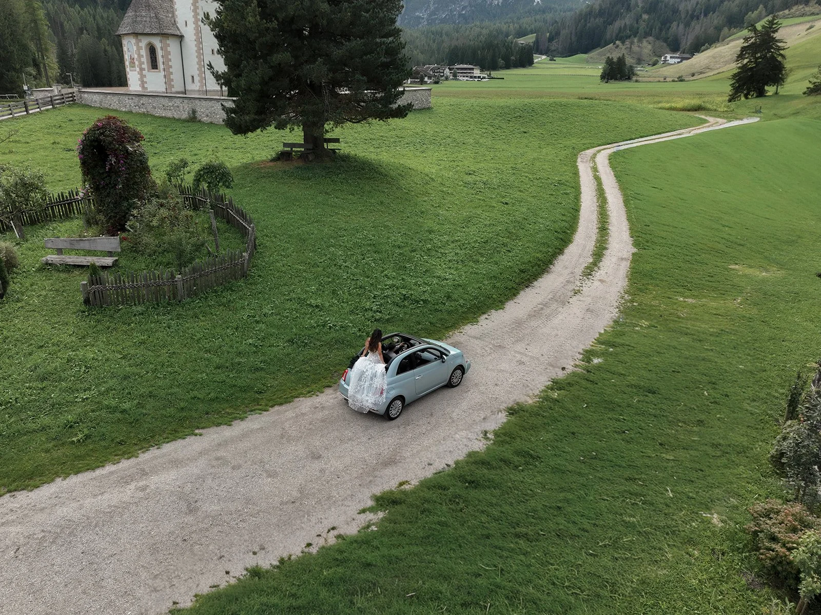 a drone photo of a bride sitting on the back of a fiat cat driving down a small country road in the dolomites in italy