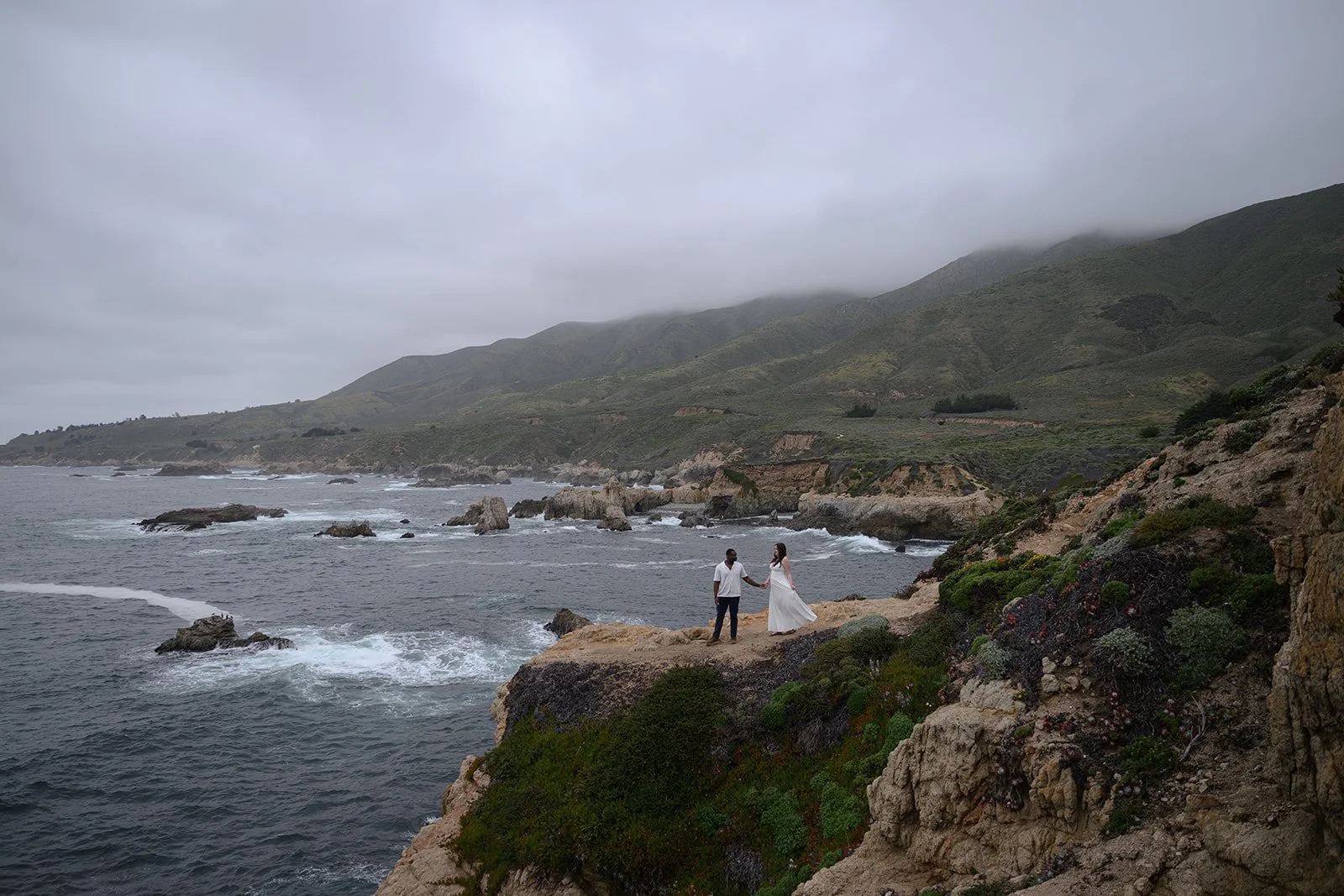 a photo of an engaged couple standing on a cliffside in big sur holding hands on a cloudy day with the rocky coastline stretching out behind them
