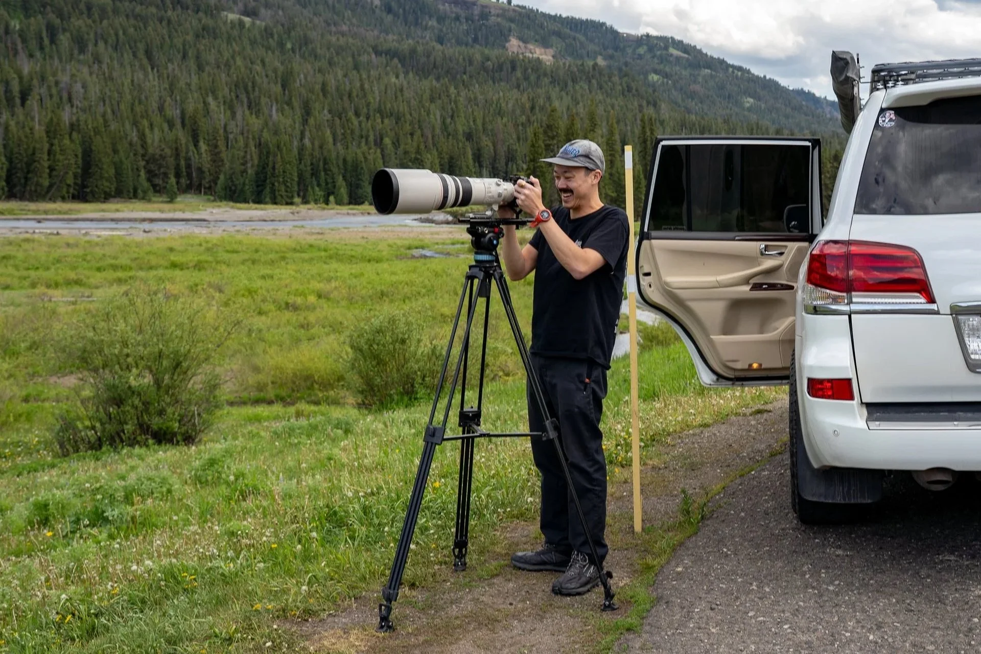 A man with a gray cap and black t-shirt using a spotting scope on a tripod outdoors next to a white vehicle with an open door, in a grassy field with trees and mountains in the background.