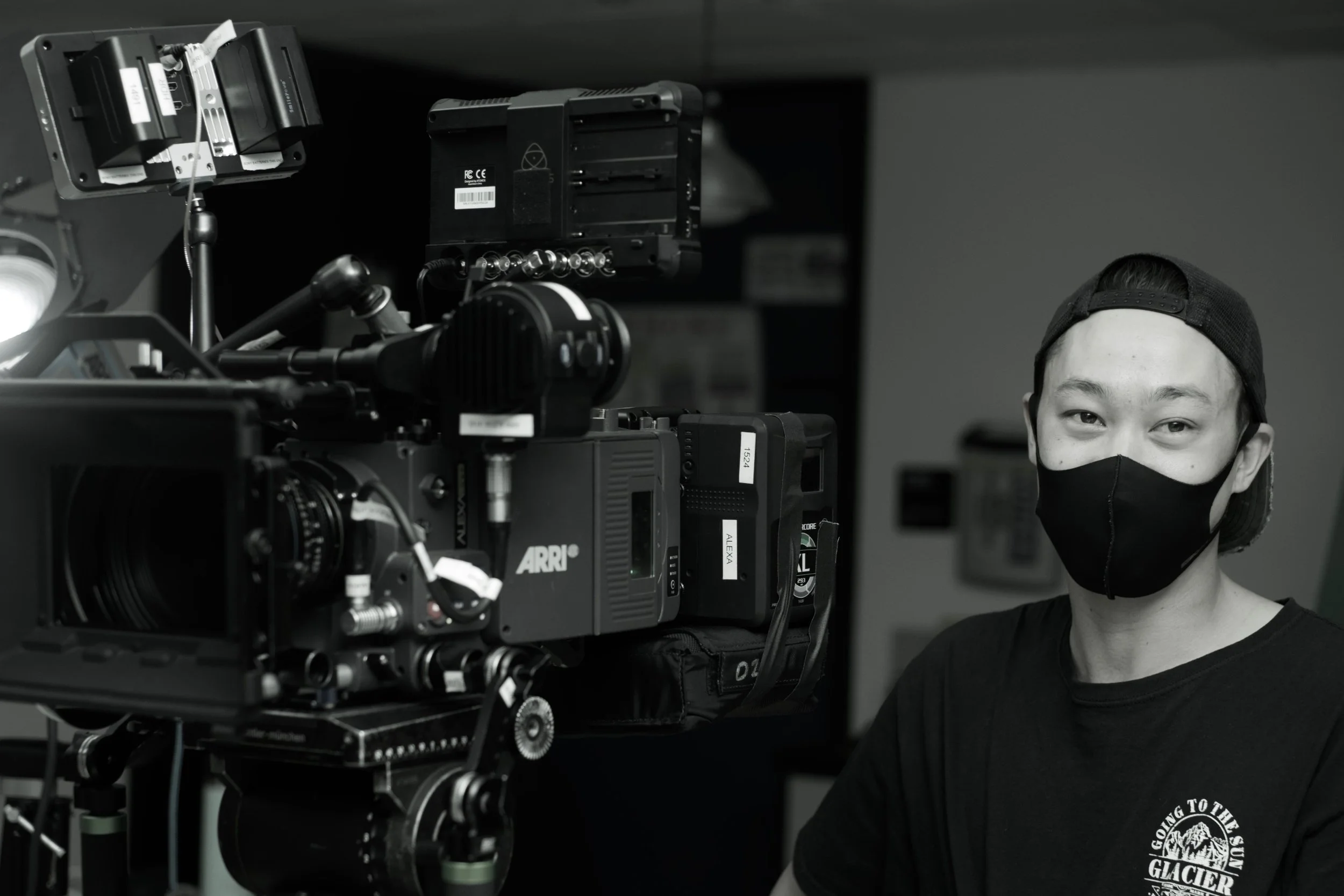 A person wearing a black face mask and a backwards baseball cap standing next to professional film equipment in a black and white photo.