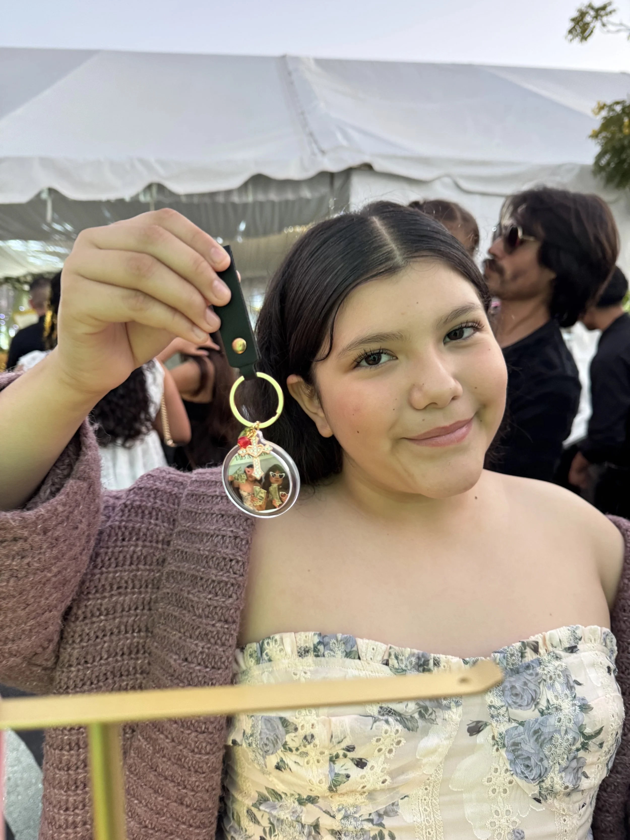 Close-up of a woman smiling and holding a keychain with a photo, at an outdoor event with people and a white tent in the background.