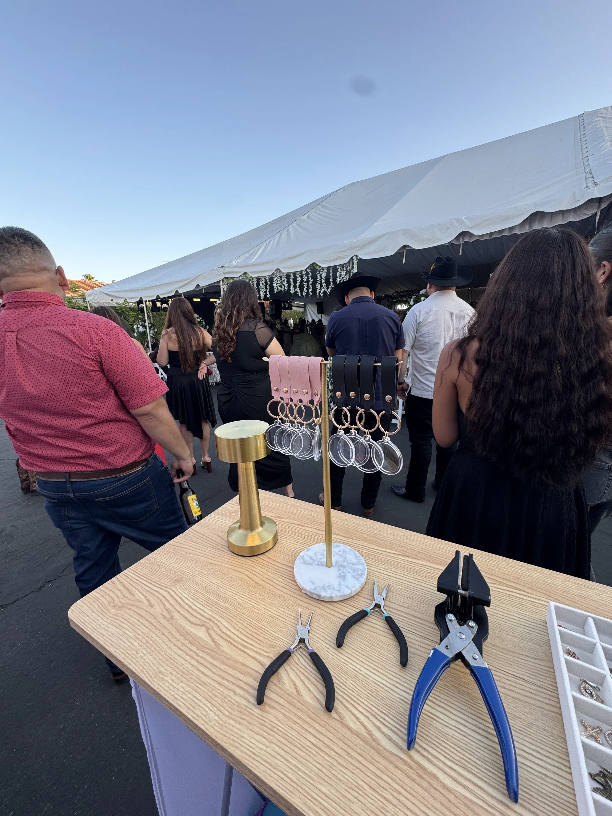 A display of jewelry-making tools including pliers and wire cutters on a wooden table, with a gold and marble jewelry stand and hanging jewelry pieces, outdoor setting with people attending an event under a white tent.