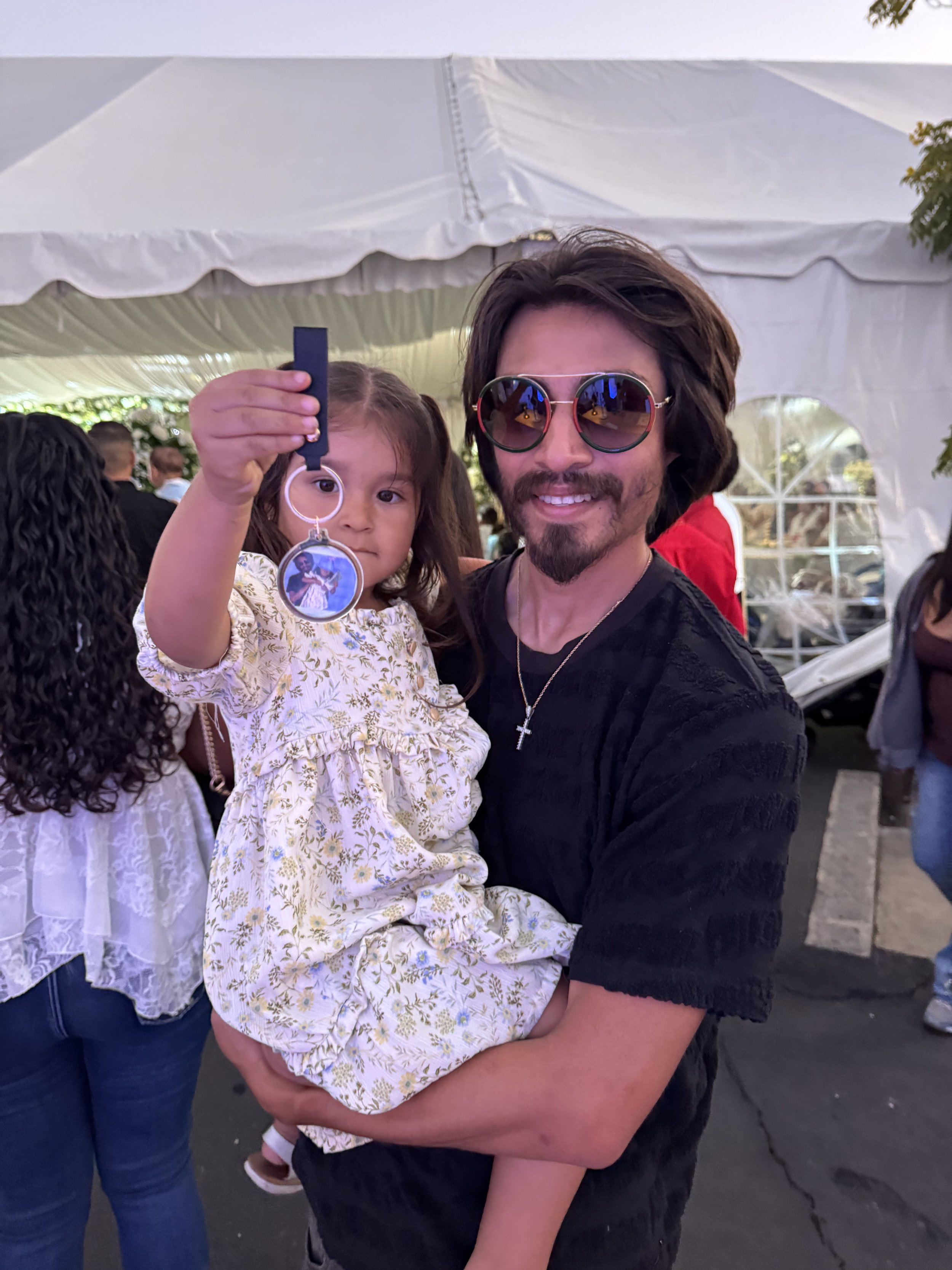 A man with shoulder-length dark hair, sunglasses, and a cross necklace holding a young girl with long brown hair in a floral dress, at an outdoor event under a white tent.