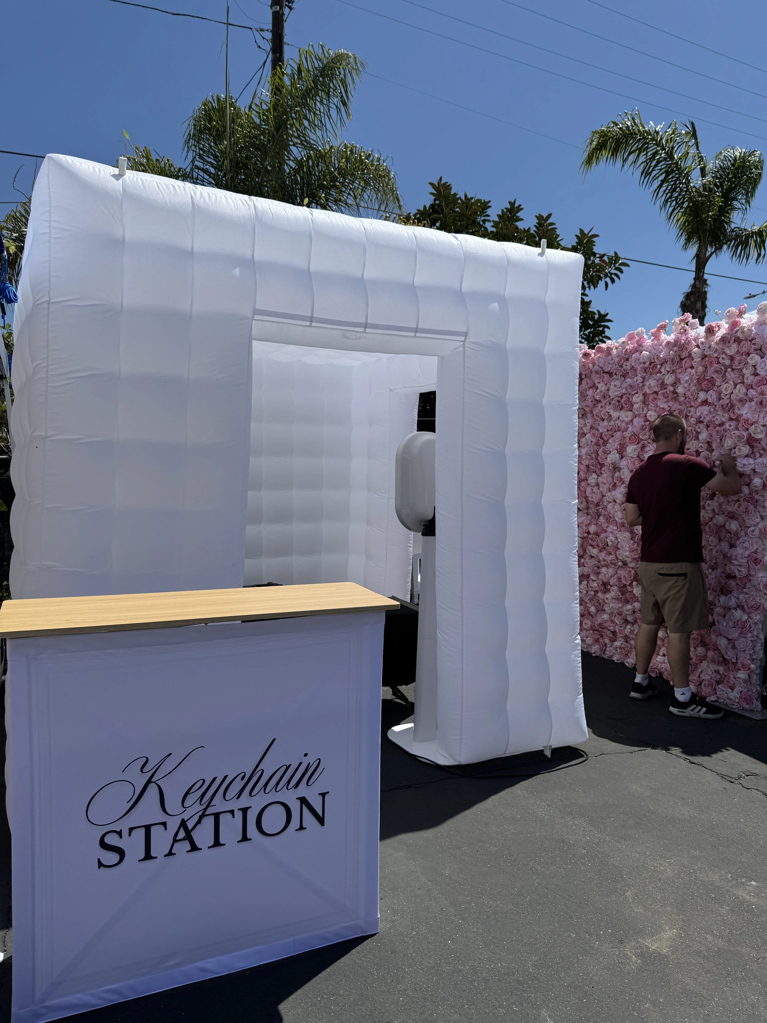 A white inflatable photo booth with the words "Keychain Station" printed on a sign in front, set up outdoors on a sunny day with palm trees and a pink rose wall in the background. A person in a burgundy shirt and khaki shorts is standing near the rose wall.