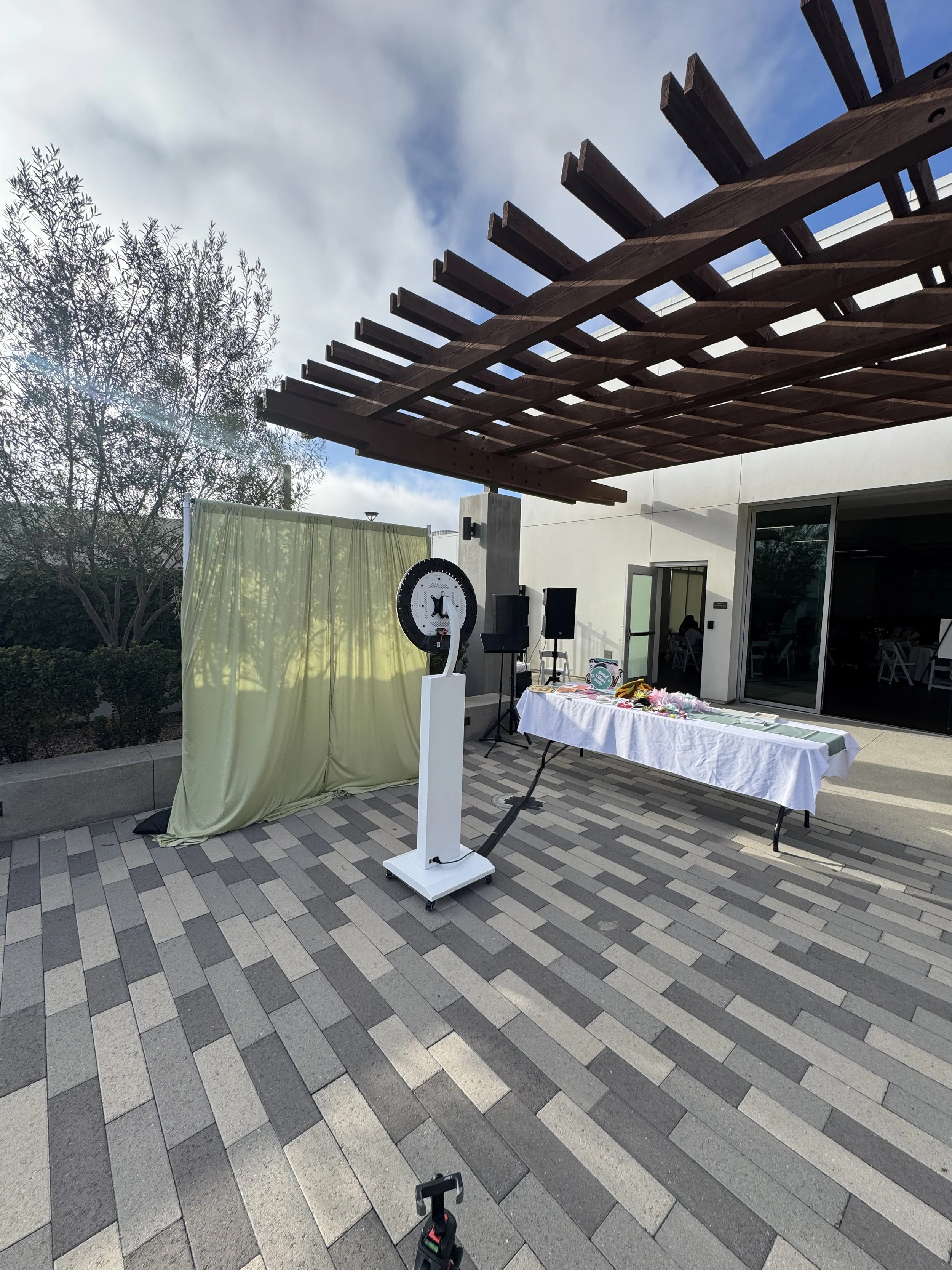 Two people smiling at a photo booth, one woman and one young man, both wearing fun hats, sunglasses, and leis, with a multicolored shiny backdrop.