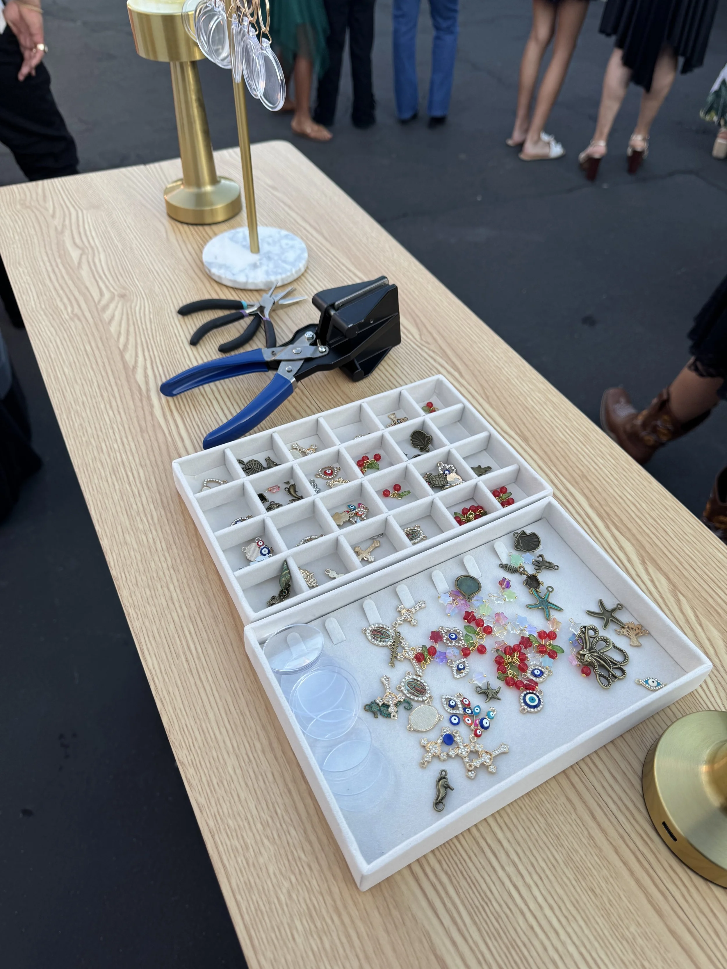 A table displaying various jewelry charms and small accessories, with a jewelry making or crafting tool, and a tray holding metal charms with decorative designs and rhinestones. In the background, people are standing at an outdoor event.