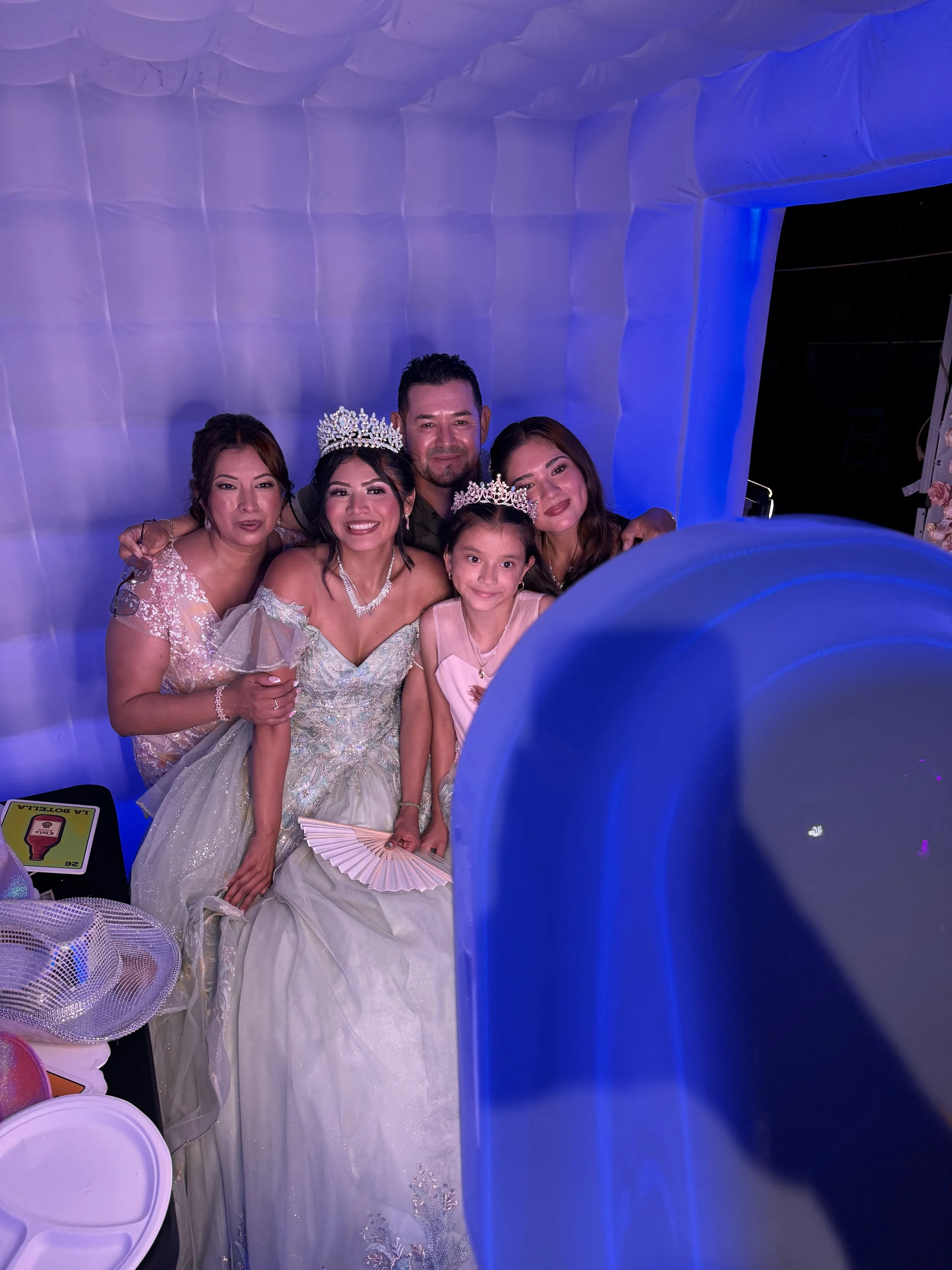 Group of five people, including children and women, celebrating a special occasion, wearing formal attire, some with tiaras and jewelry, inside a decorated venue with white and purple lighting.