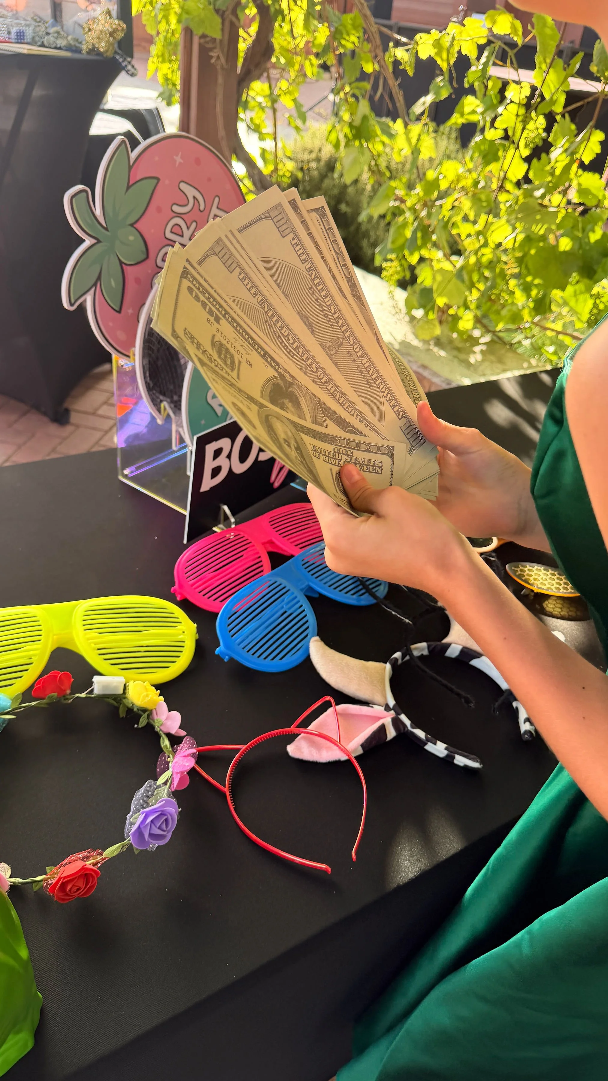 A person holding a fan of U.S. dollar bills. The table in front has colorful sunglasses, a flower headband, and some small decorative items. There is a sign in the background with a strawberry and the word 'Berry.'