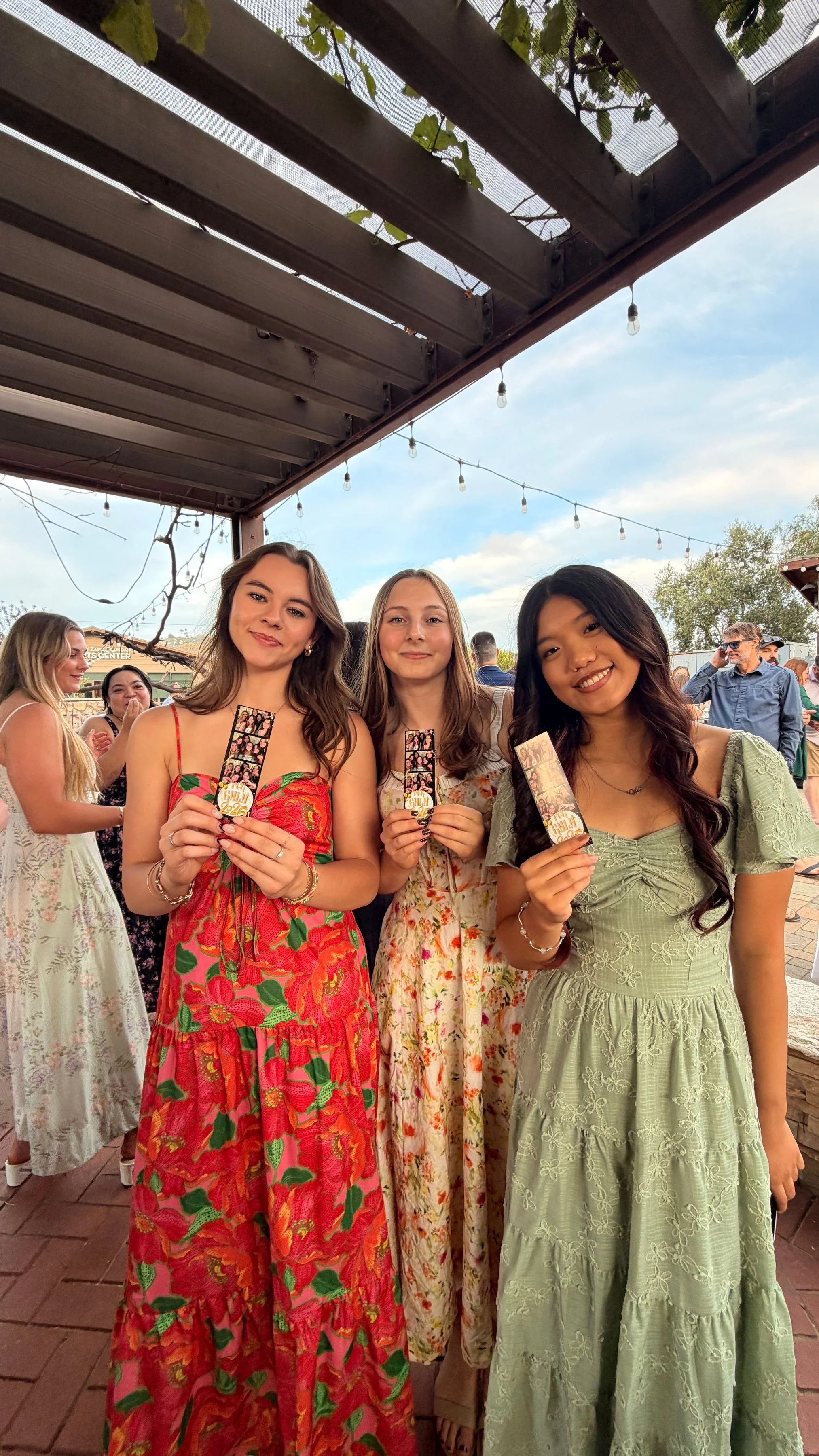 Three young women are standing together at a celebration, holding photo booth props, wearing colorful dresses, and smiling at the camera. The setting is outdoors with string lights and other people in the background.