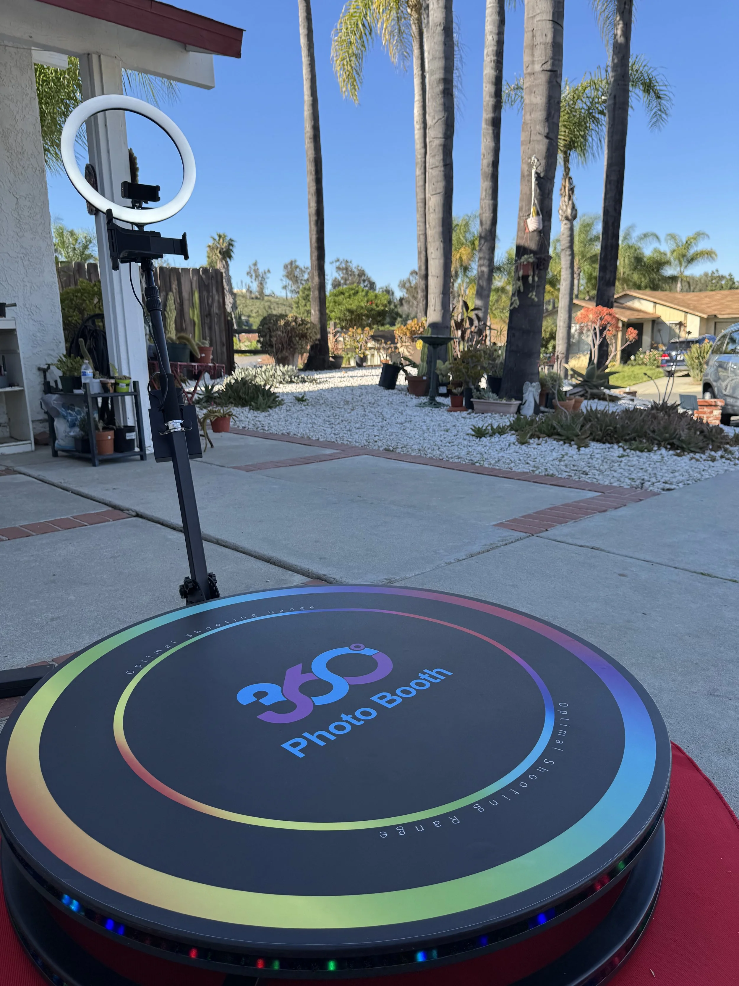 A circular photo booth with a colorful border and the words 'Photo Booth' on the top, situated outdoors on a concrete patio with a house, trees, and blue sky in the background.