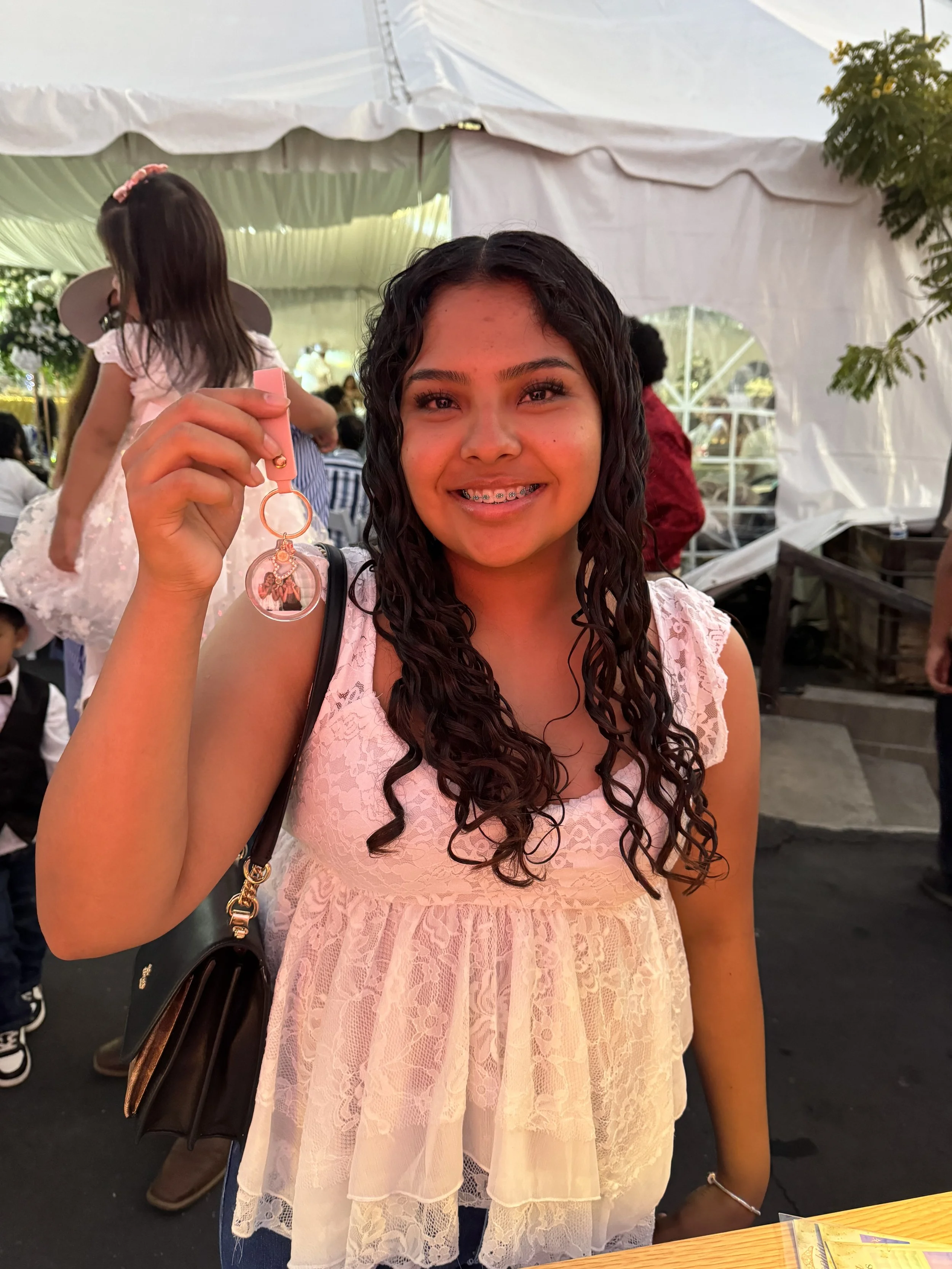 A smiling young woman with curly dark hair and braces holding a round keychain with a photo, standing indoors at an event with a white tent and other people in the background. She is wearing a pink lace dress and a bracelet on her right wrist.