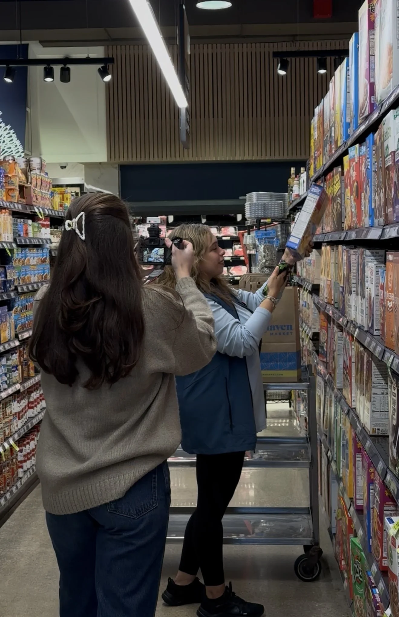 Two women shopping in a grocery store aisle, one taking a photo of the other who is examining cereal boxes.
