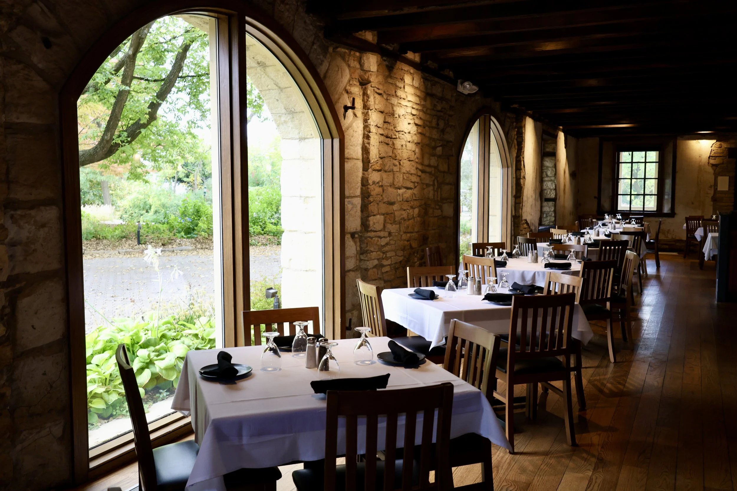 Interior of a restaurant with large arched windows, wooden beams, stone walls, and tables set with white tablecloths, black napkins, and glassware.