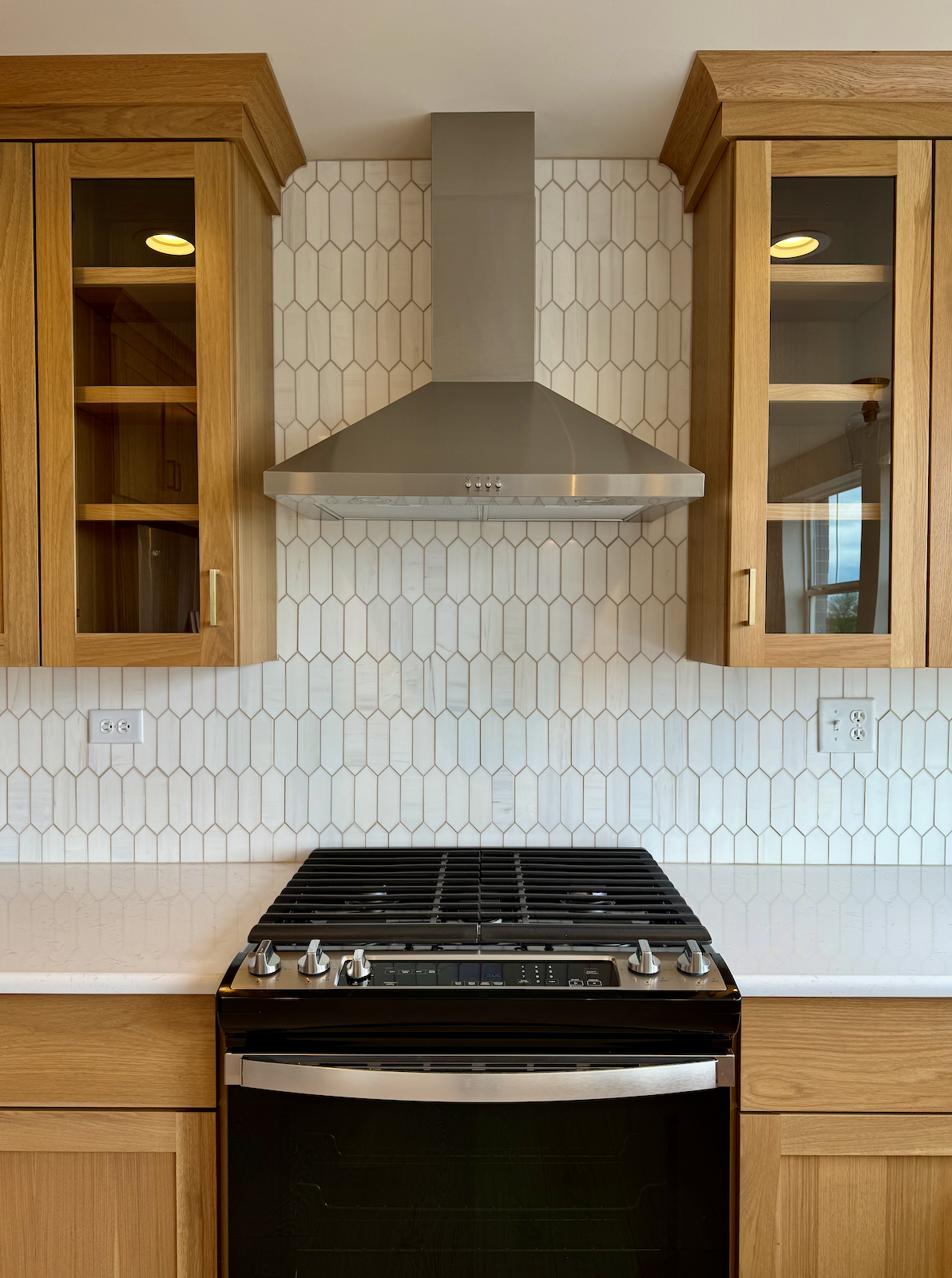 A modern kitchen with a stainless steel range hood above a black stove, wooden cabinets on either side, and a backsplash of white hexagonal tiles.