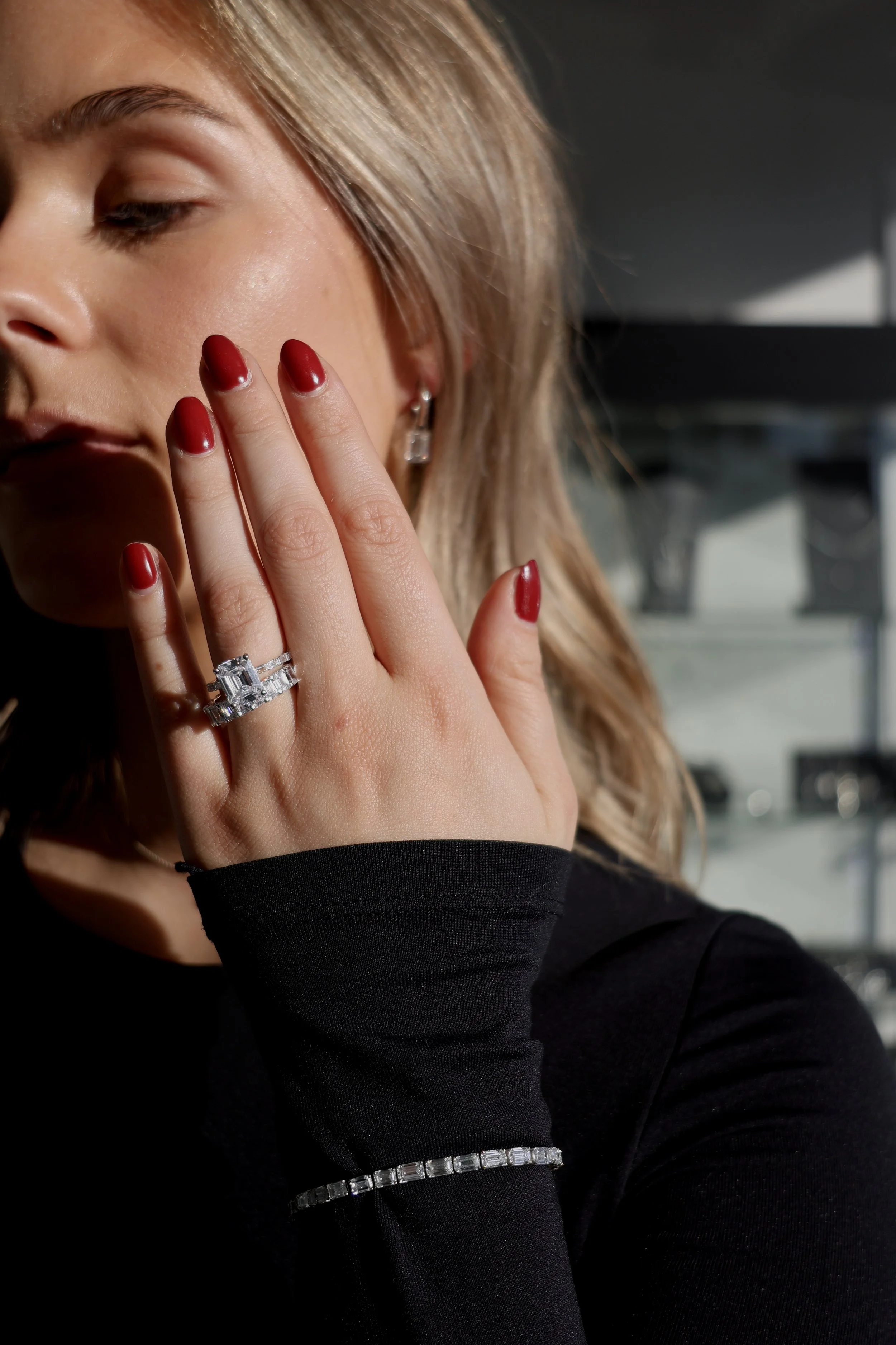 Close-up of a woman with blonde hair and red nails, showing a silver ring with a large rectangular gemstone, wearing a black long sleeve shirt and a matching bracelet.