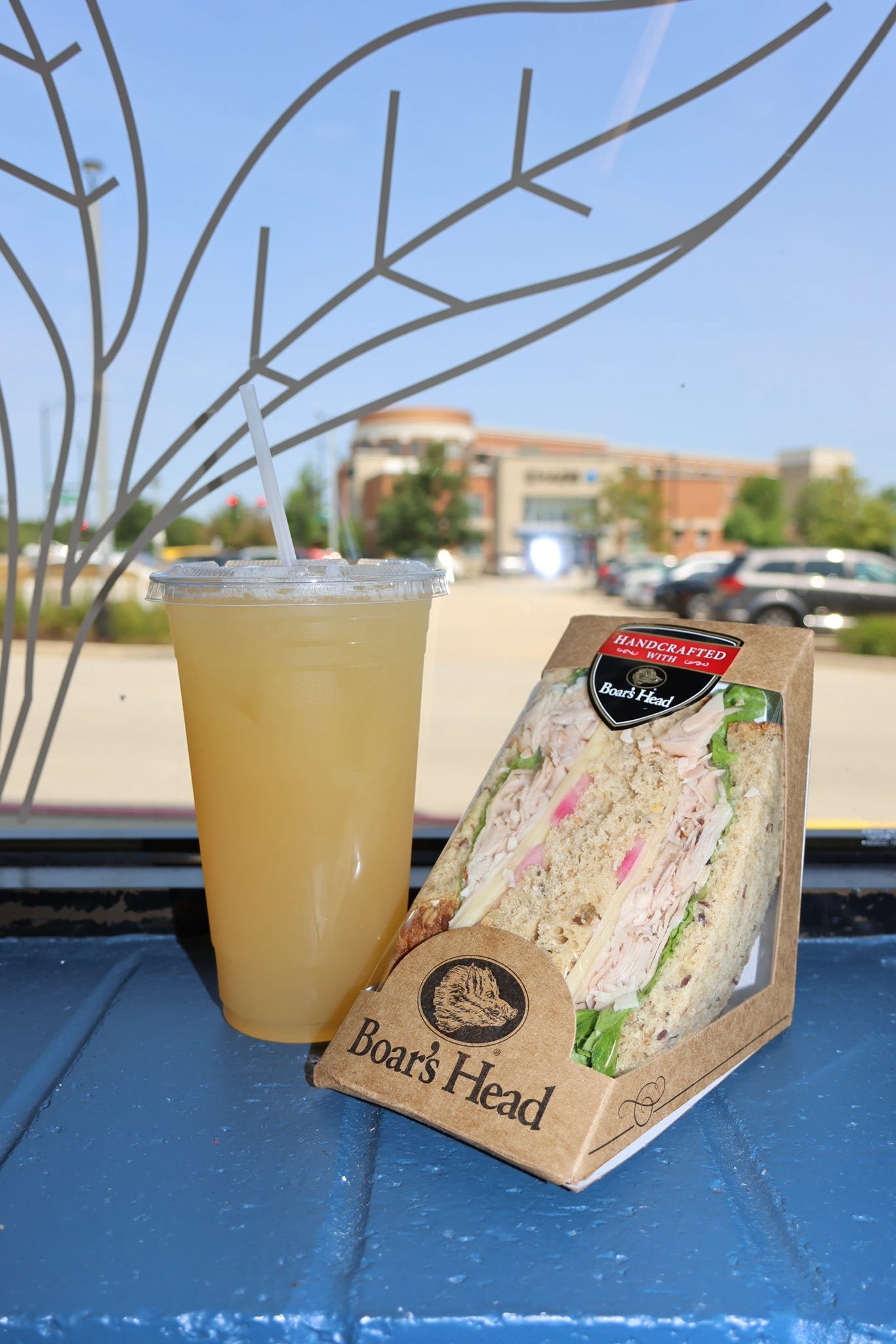 A sandwich in Boar's Head packaging and a yellow beverage with a straw, placed on a blue surface outside with a parking lot and shopping mall in the background.