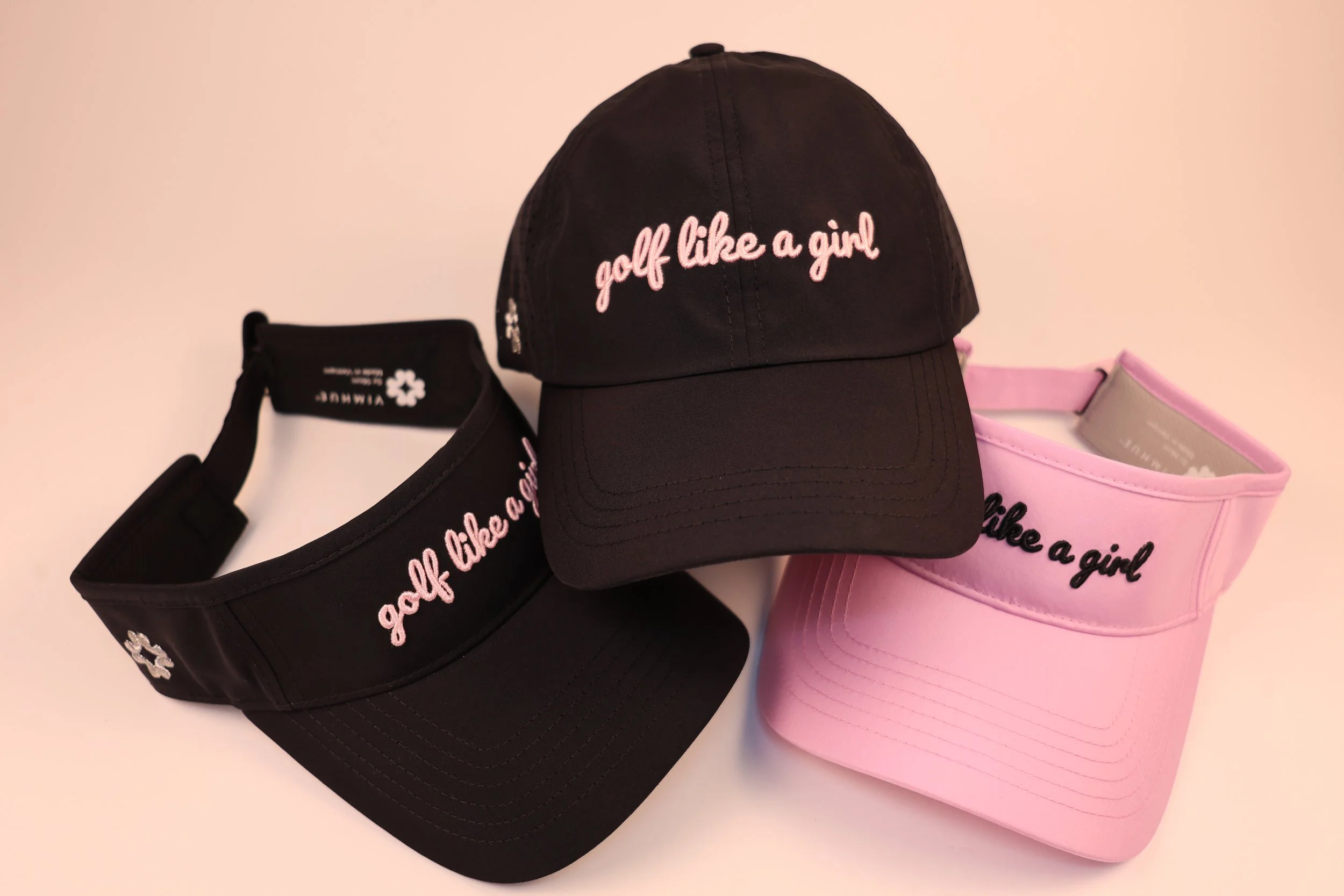 Black and pink golf visors and a black baseball cap, all with embroidery that says 'golf like a girl'.