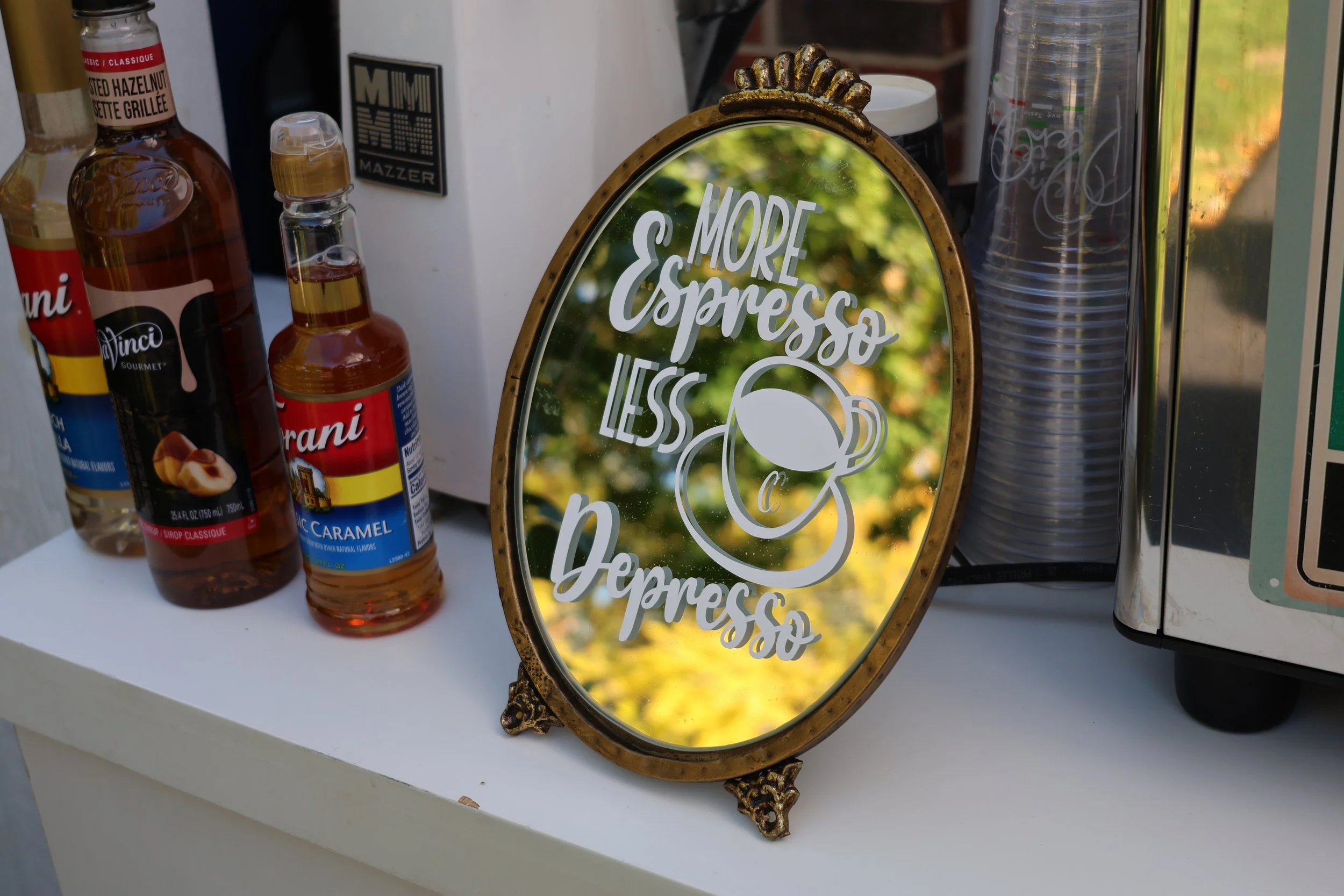 A vintage oval mirror with a brown frame displaying the phrase 'More Espresso Less Depresso' and an illustration of a coffee cup, placed on a white surface next to syrup bottles, a stack of cups, and a metal appliance.