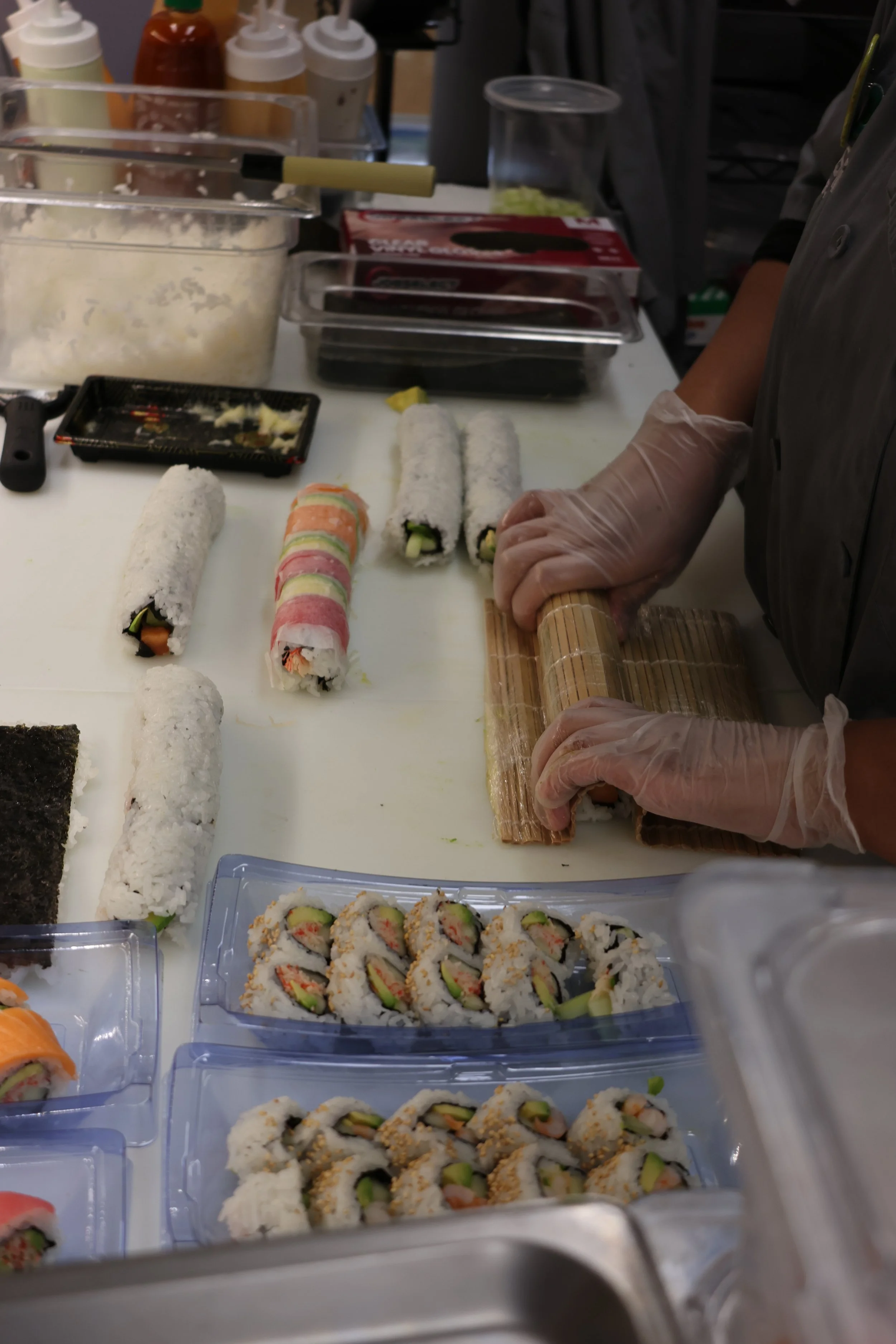A person wearing gloves is rolling sushi with a bamboo mat on a white work surface, with various sushi rolls and ingredients around.