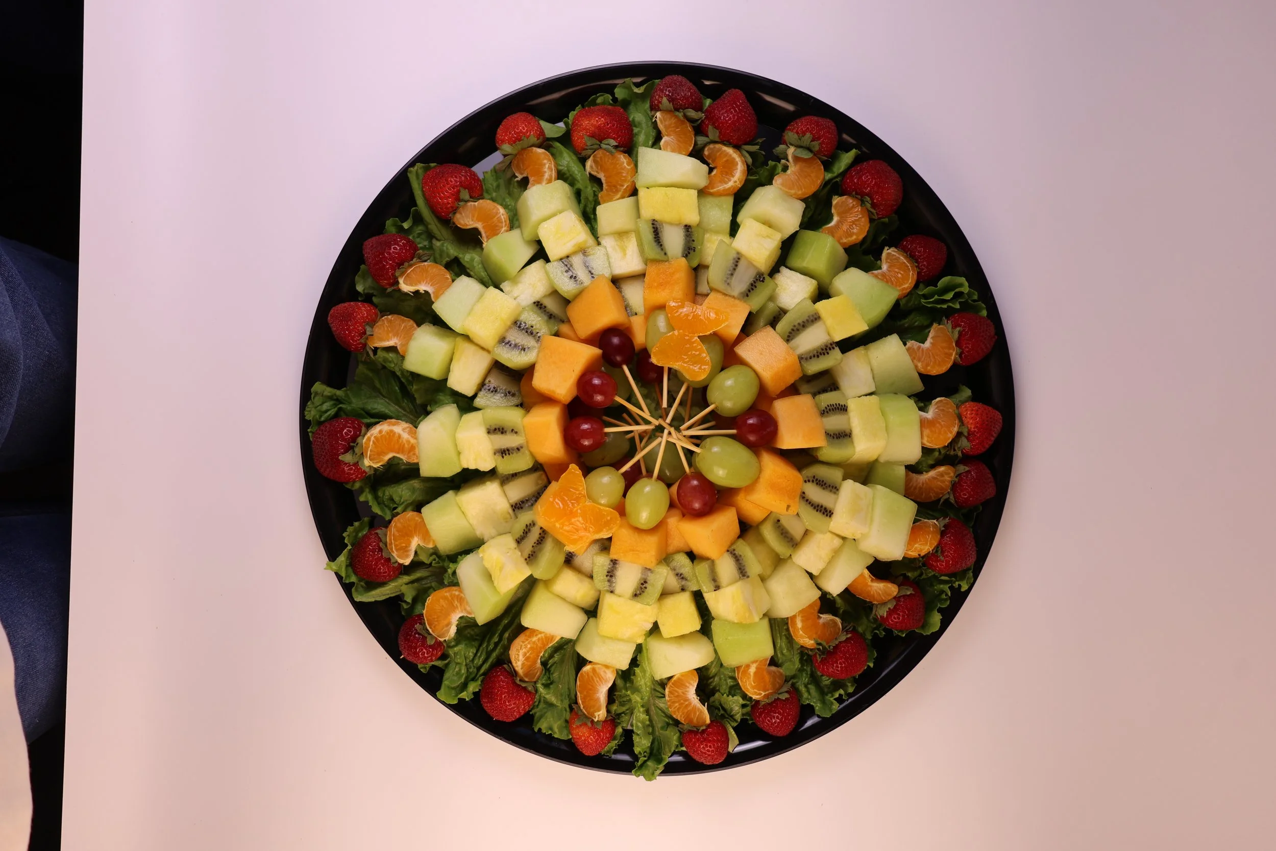 A large round platter of assorted fresh fruit arranged in concentric circles with strawberries, tangerine slices, honeydew, cantaloupe, kiwifruit, grapes, and leafy greens.