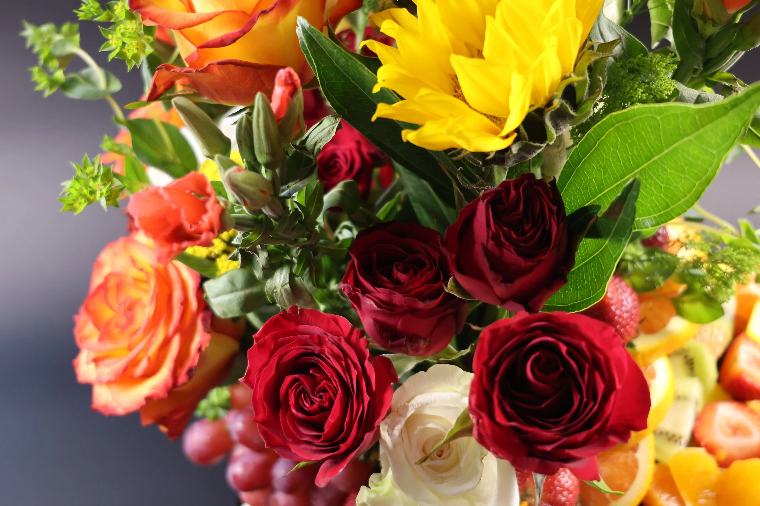 Close-up of a floral arrangement with red, white, and orange roses, yellow sunflower, green leaves, and other assorted flowers and fruits.