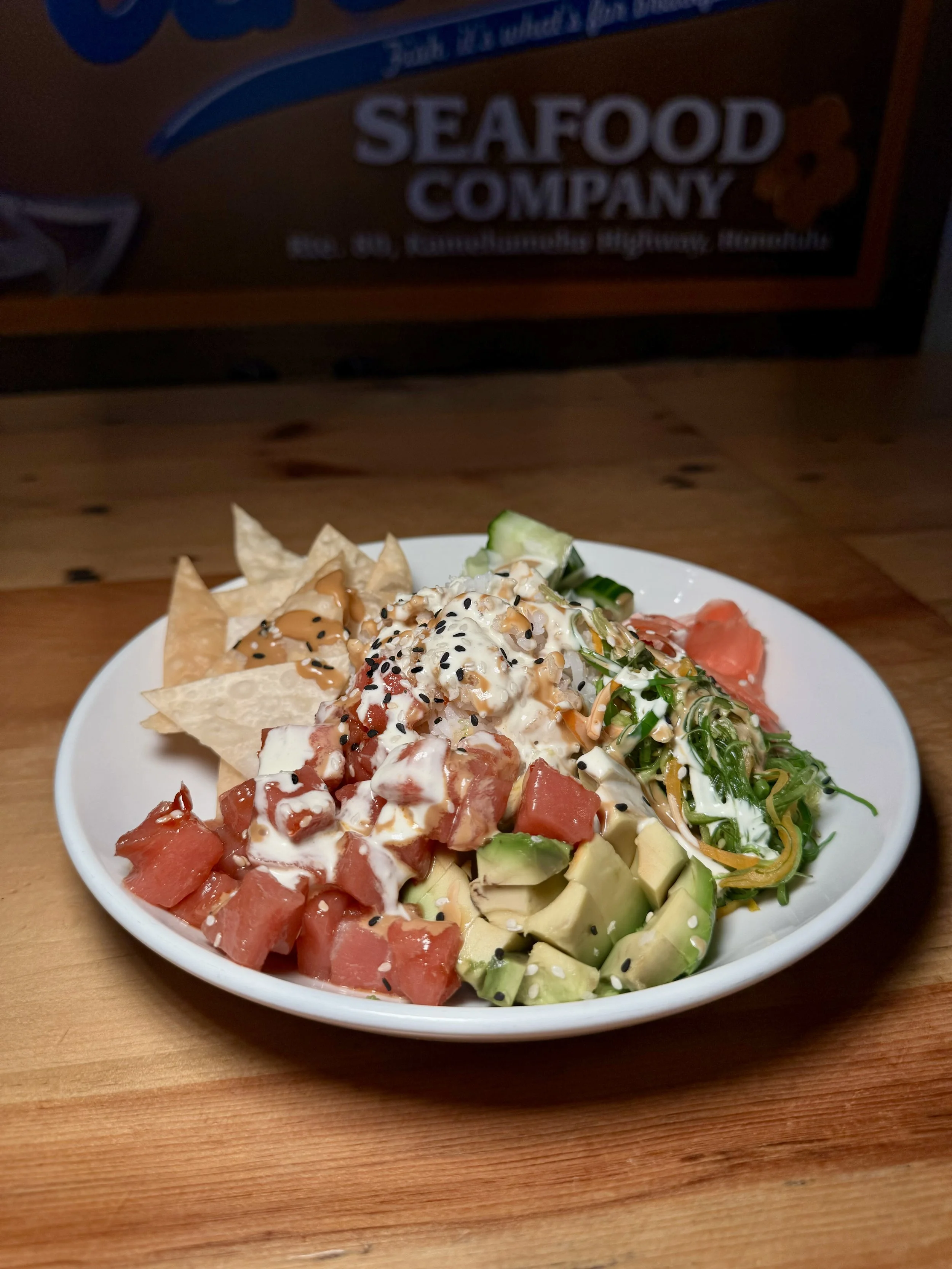 A bowl of assorted poke ingredients including chopped watermelon, avocado, cucumber, shredded vegetables, and diced raw fish topped with sauce, sesame seeds, and black sesame seeds, with tortilla chips on the side.