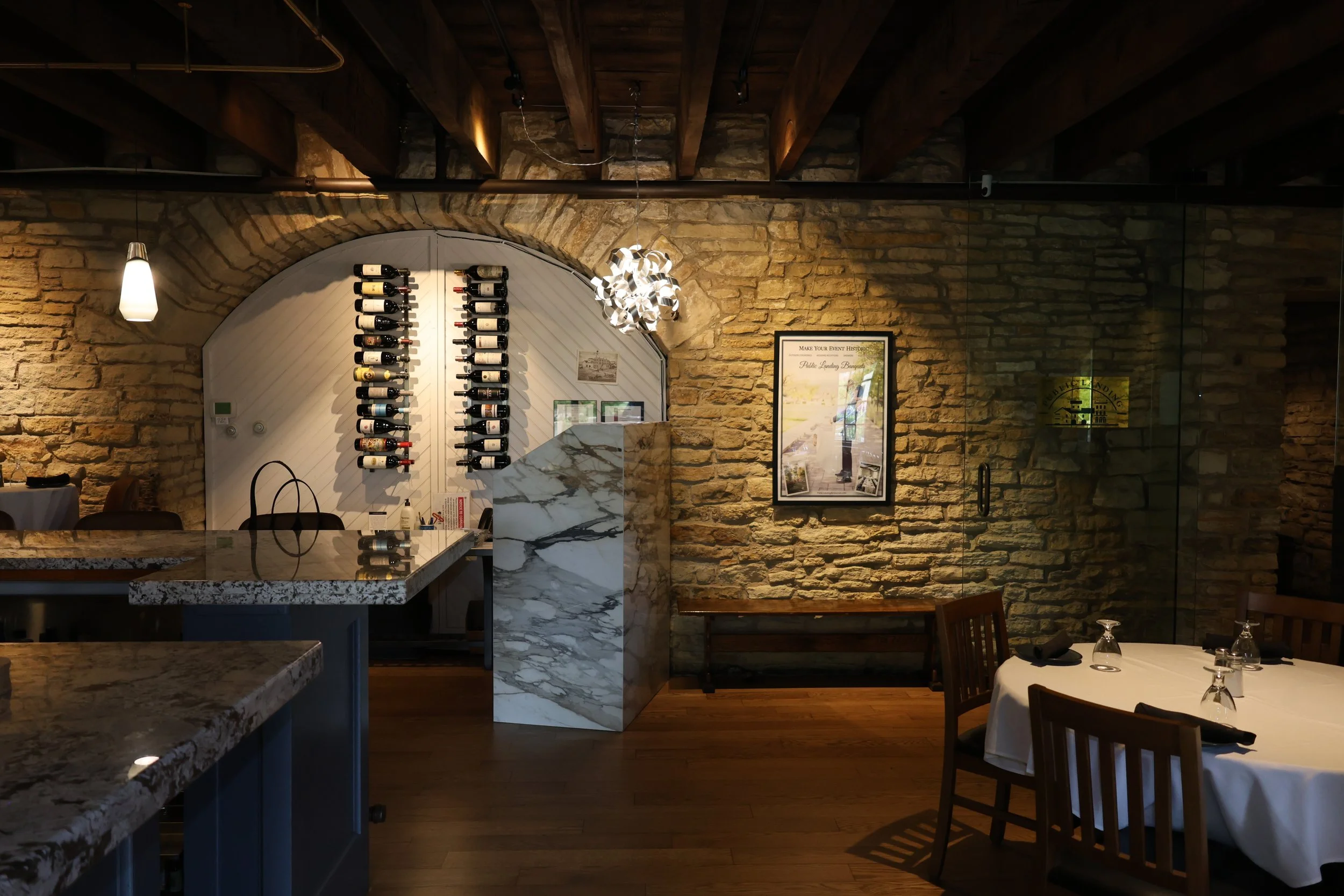 Interior of a restaurant with exposed brick walls, a wine display, and a round table set with glassware and napkins.