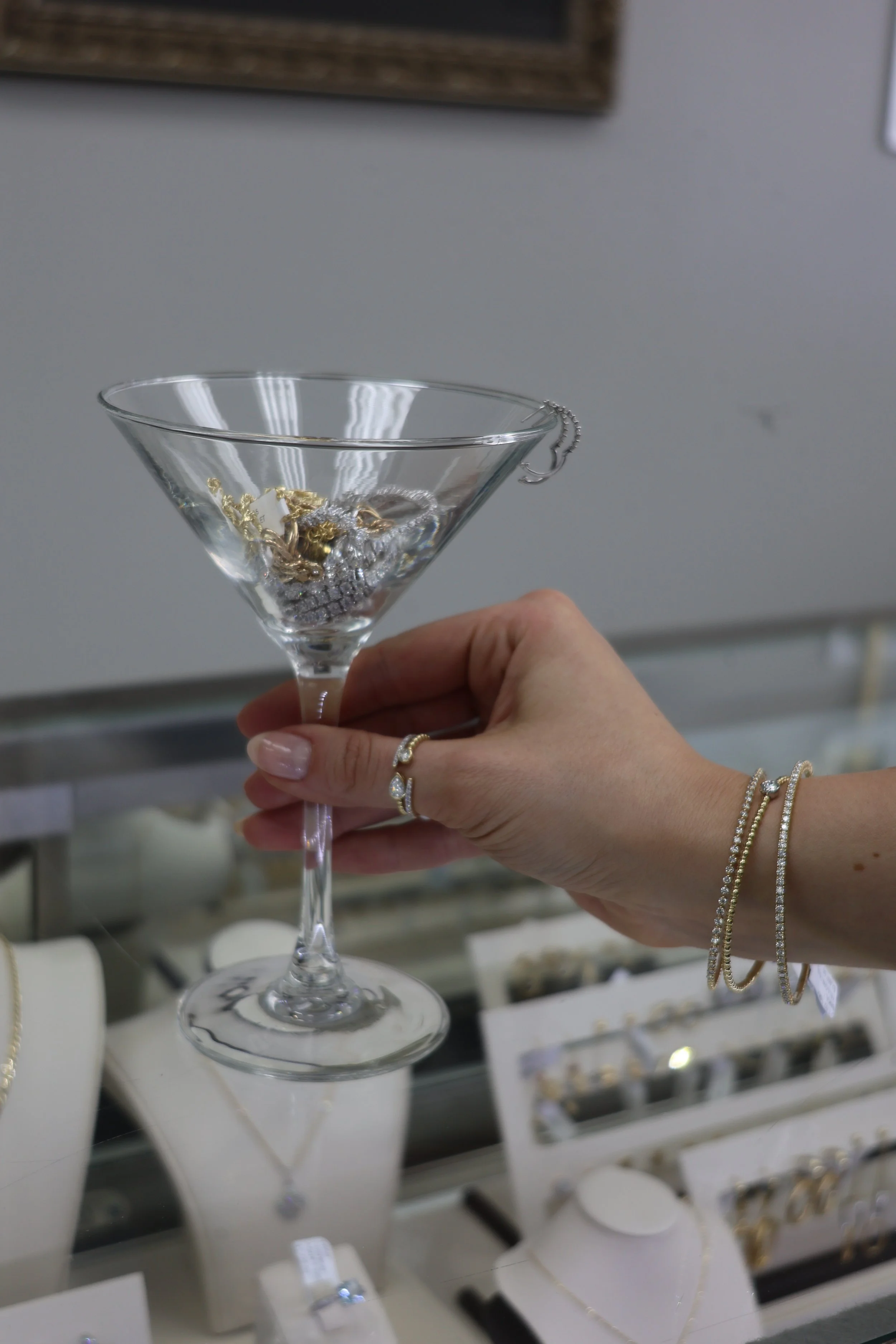 Close-up of a hand holding a glass bowl filled with jewelry, including necklaces and bracelets, with a display case of jewelry in the background.