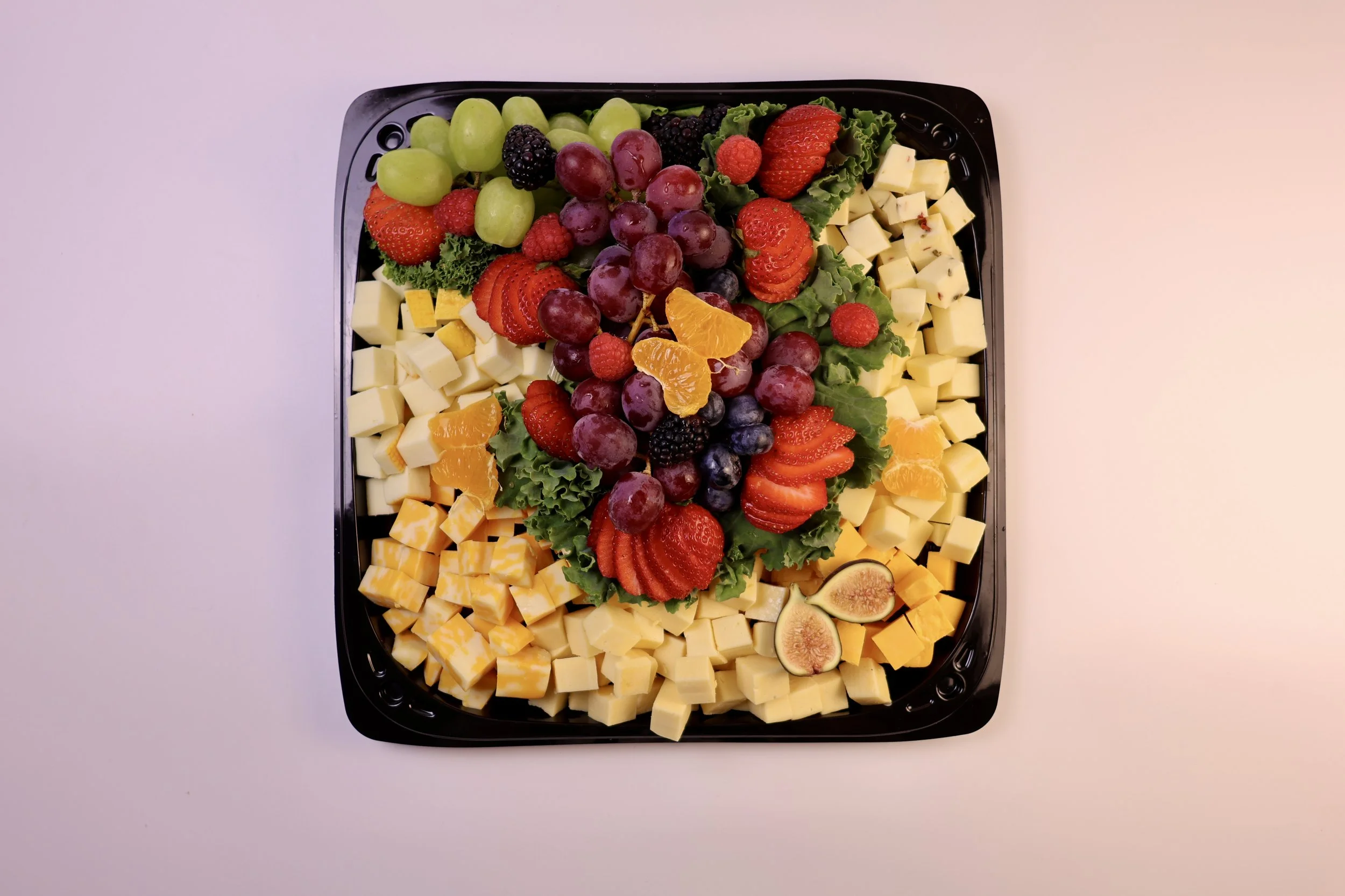 A tray of assorted cheeses, grapes, and fresh fruit including strawberries, strawberries, figs, oranges, and blackberries, arranged on a black platter with greens.