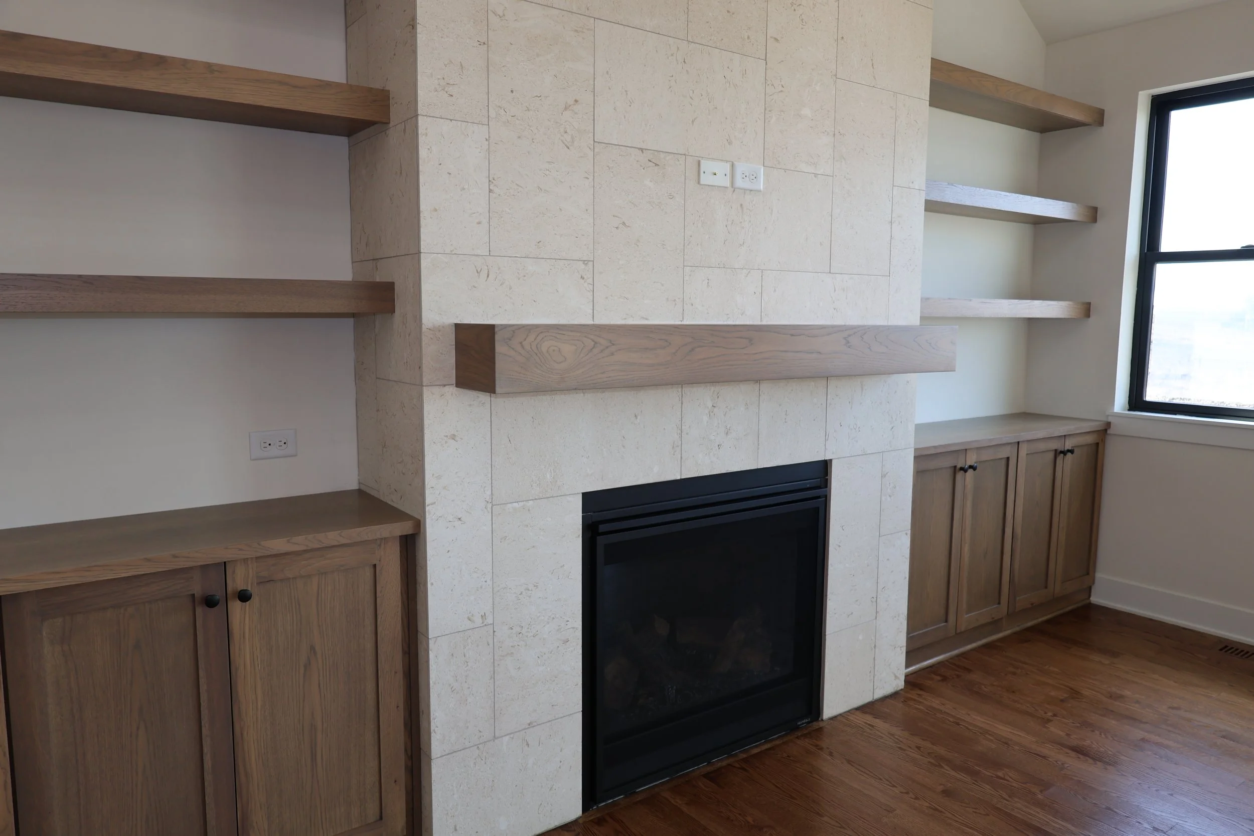 Living room corner with a fireplace, wooden shelves above and to the side, and a wooden cabinet below the shelves. A window on the right side.
