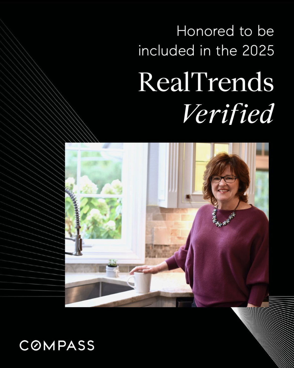 A woman in a maroon sweater and glasses standing in a bright kitchen with a window, holding a coffee mug, smiling.