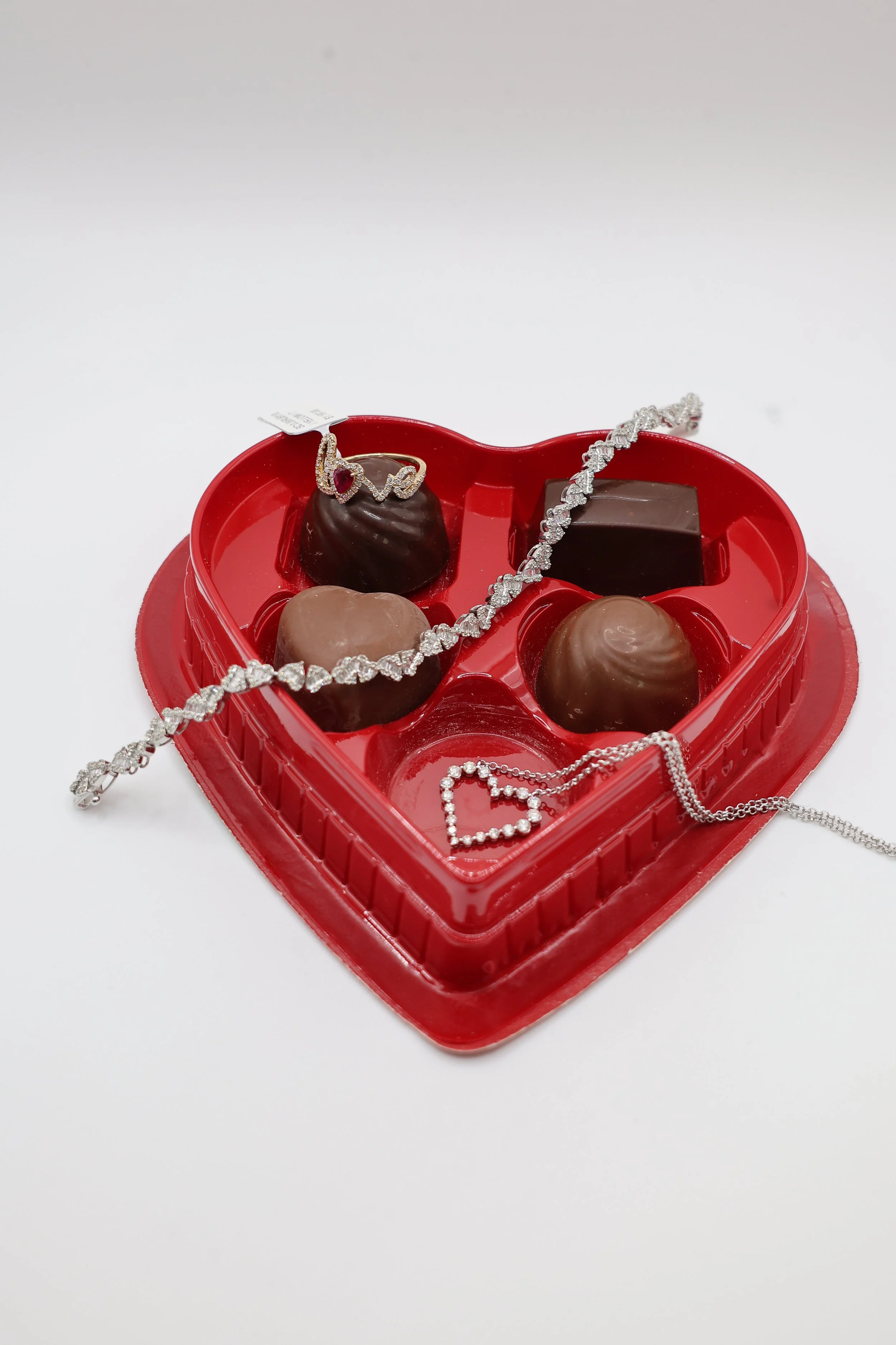 A heart-shaped box of assorted chocolates with jewelry, including a tiara, necklace, and bracelet, on a white surface.