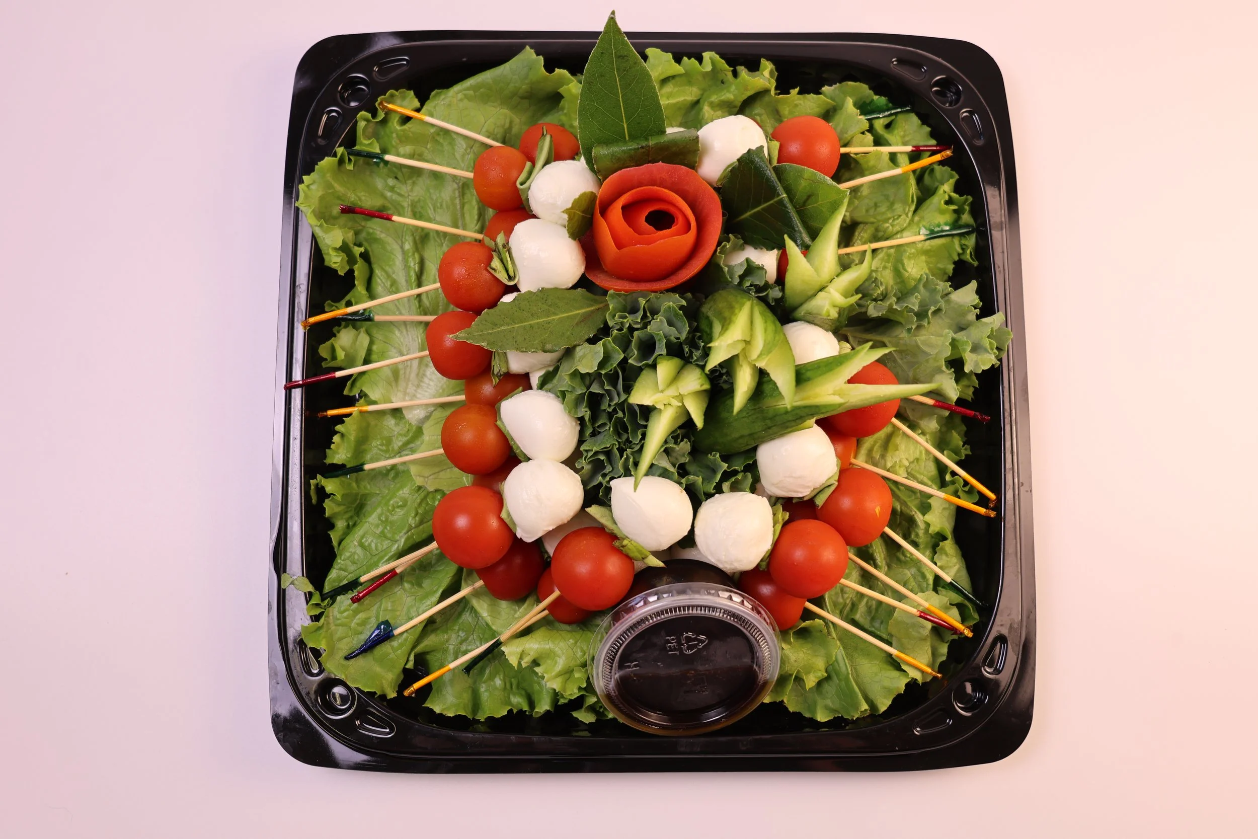 A platter of arranged vegetables and fruits, including cherry tomatoes, mozzarella balls, cucumber slices, lettuce, and decorative leaves, with a soy sauce dish in the center.