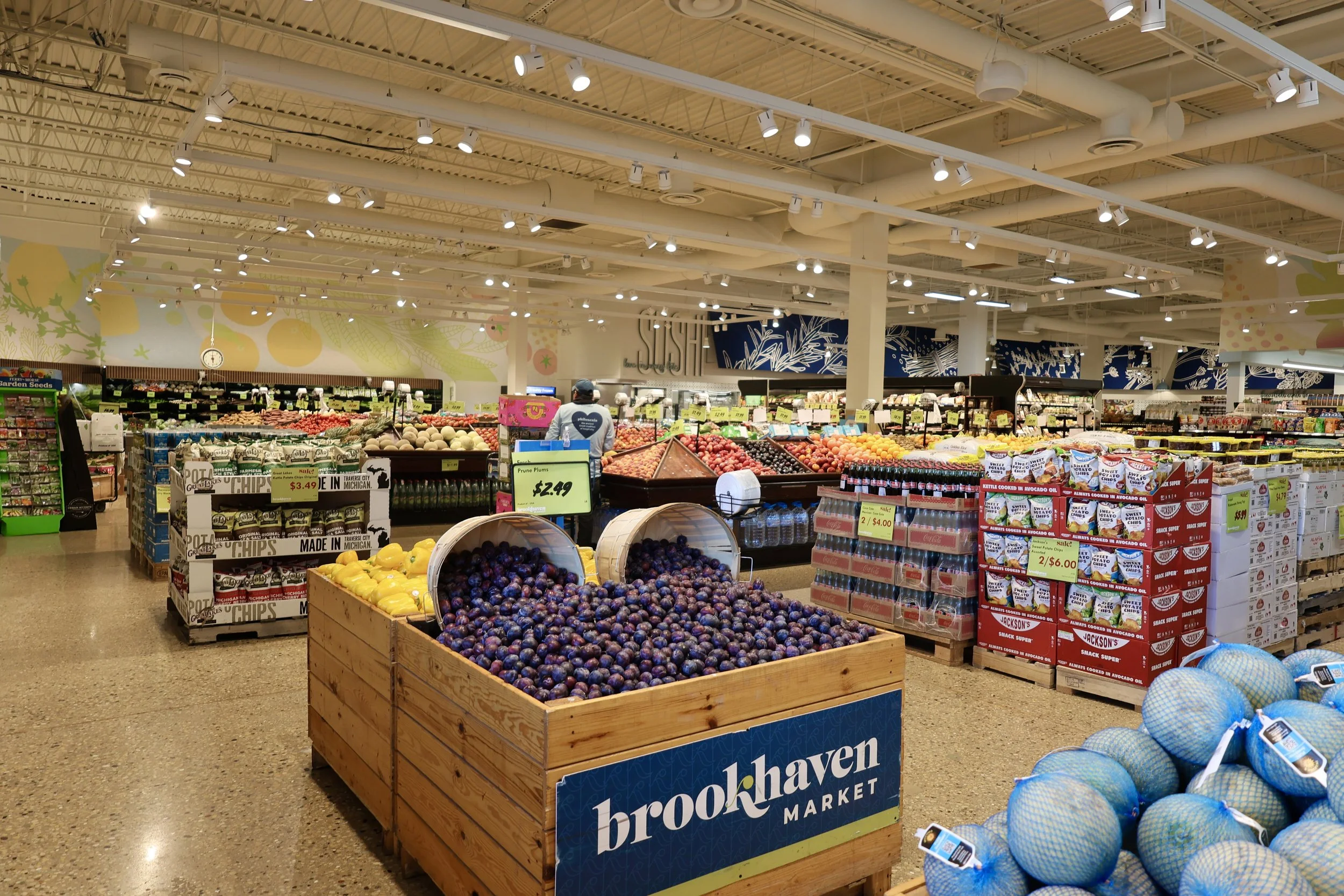Inside a grocery store with fresh produce including blueberries, plums, and yellow peppers displayed in wooden bins and baskets, with shelves and signs indicating prices.