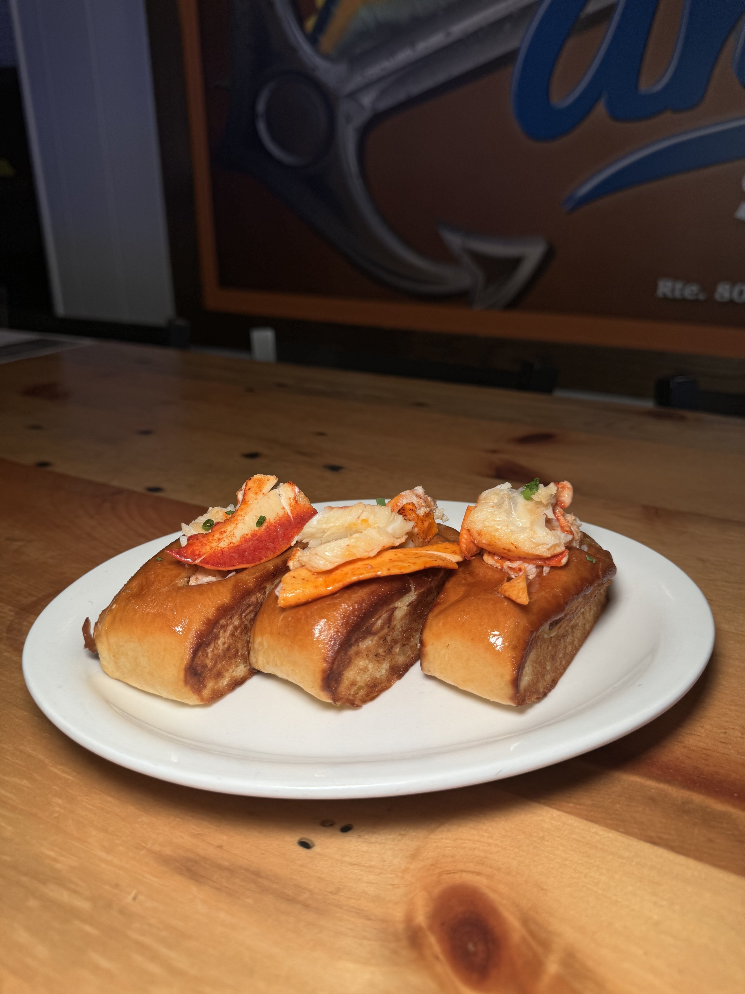 Three hot dogs topped with lobster meat on a white plate, placed on a wooden table.