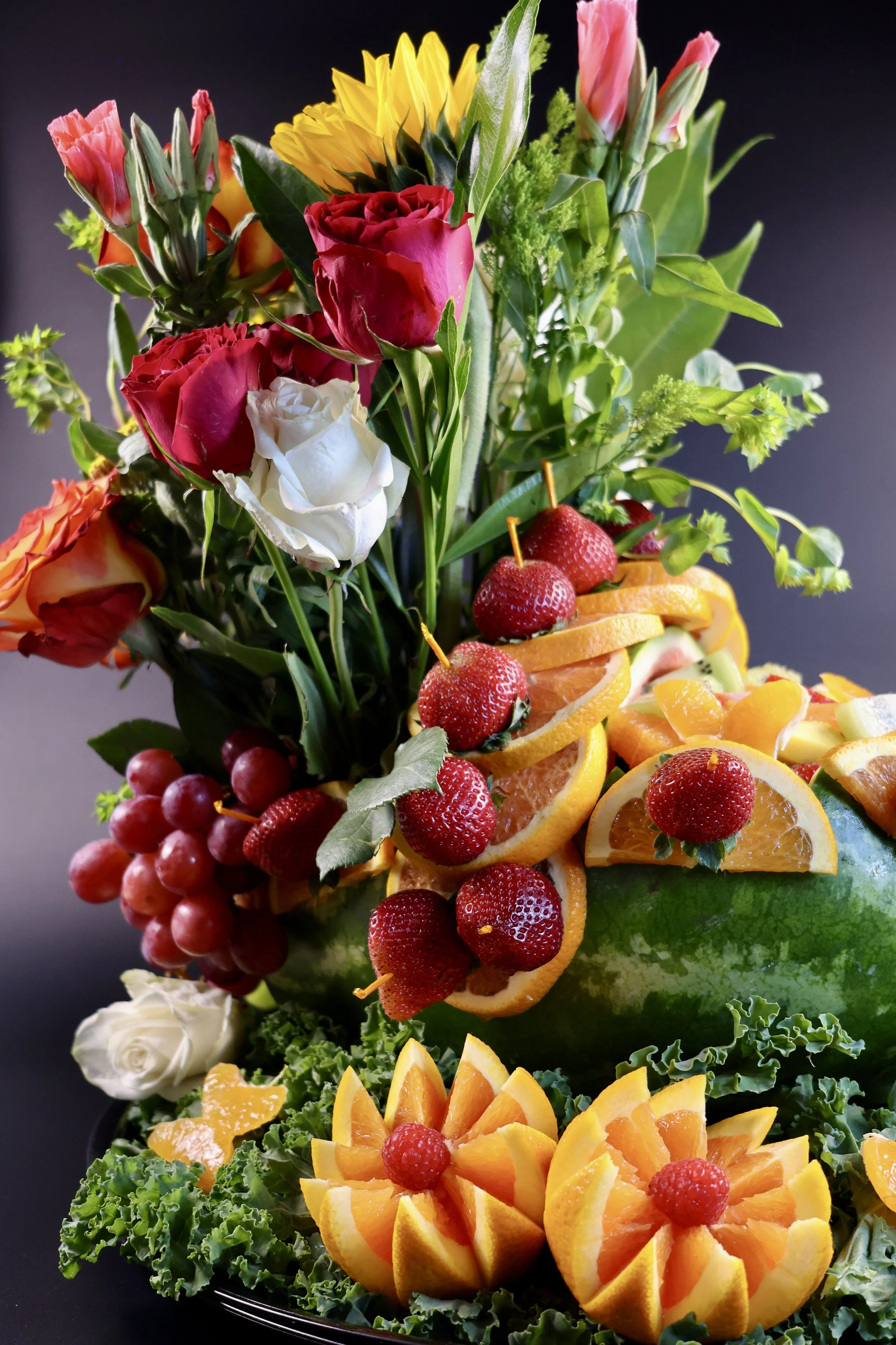 A floral arrangement with various flowers and fruits, including roses, sunflowers, strawberries, and orange slices, all displayed on a black background.