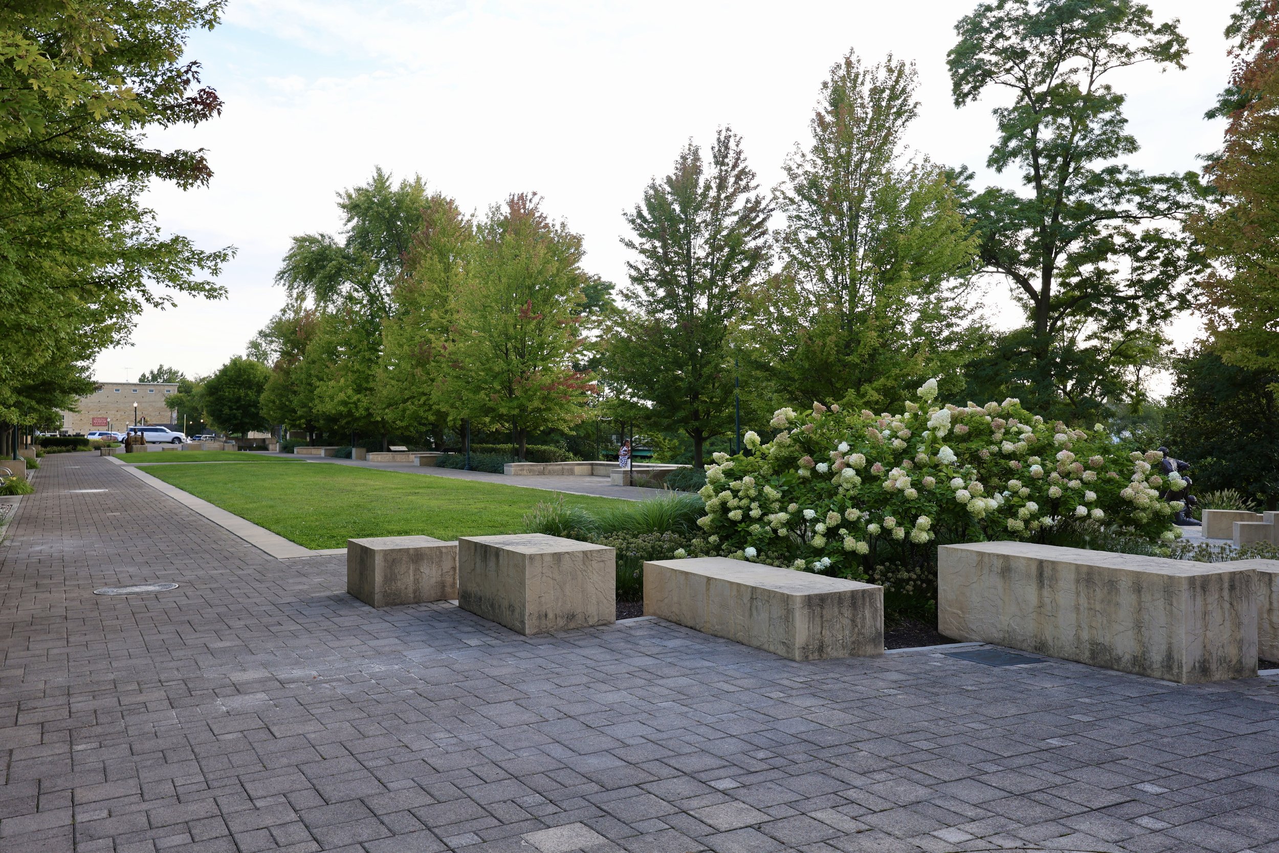 A park with a paved walkway, a grassy area, trees, shrubs with white flowers, and concrete benches under a cloudy sky.