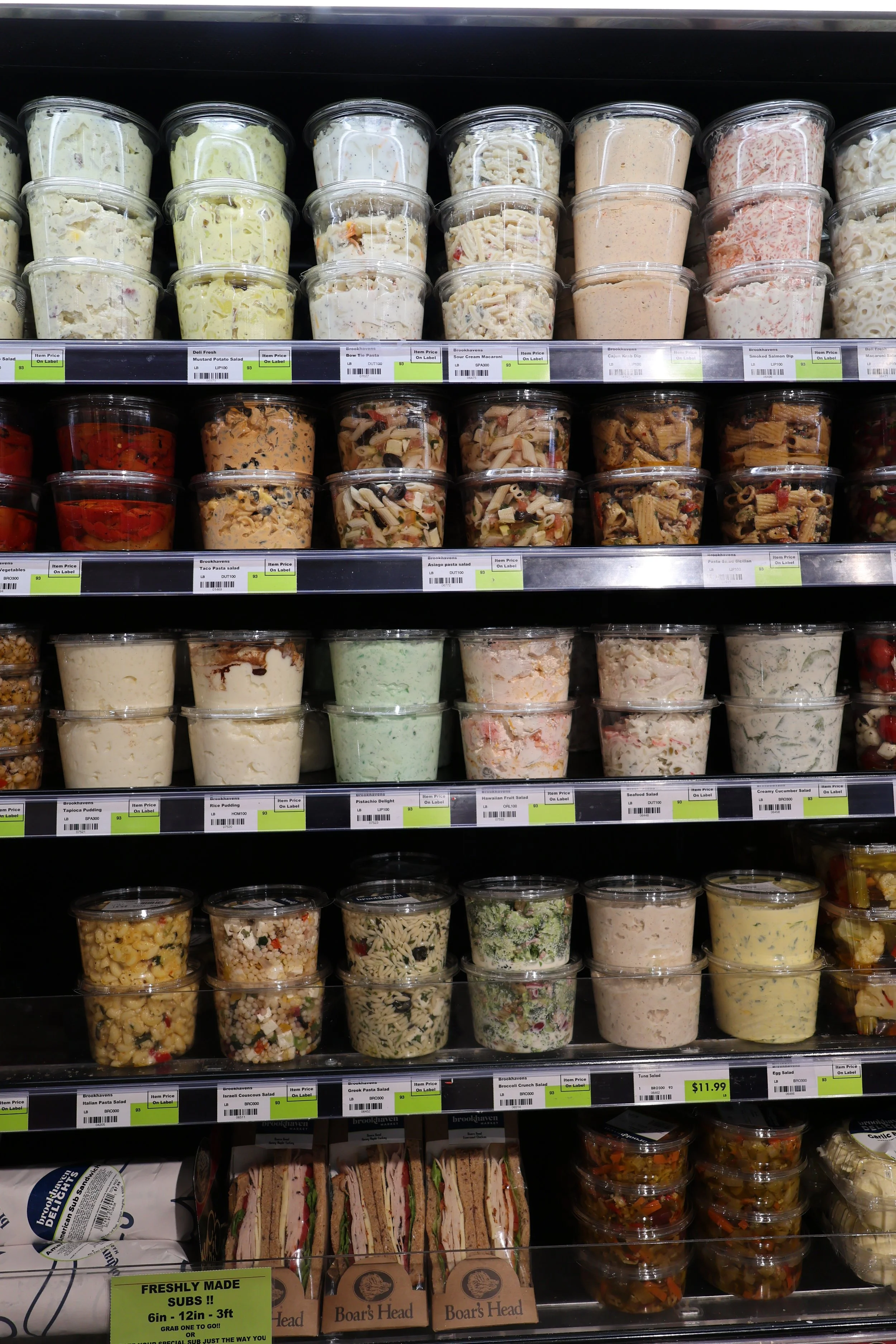 A grocery store shelf stocked with various pre-packaged salads, pasta salads, and deli sandwiches.