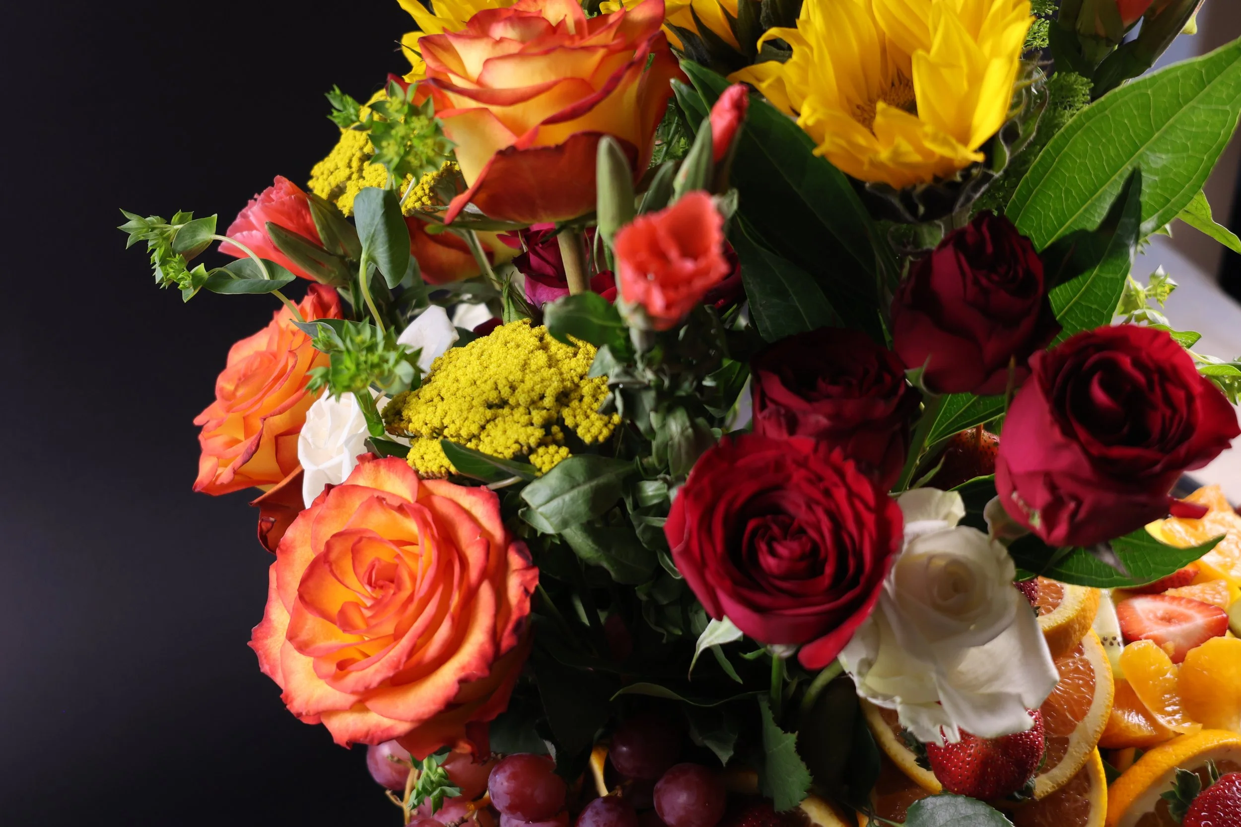 A bouquet of colorful flowers including roses, sunflowers, and other blooms with various shades of red, orange, yellow, and white, along with green foliage, contrasted against a dark background.