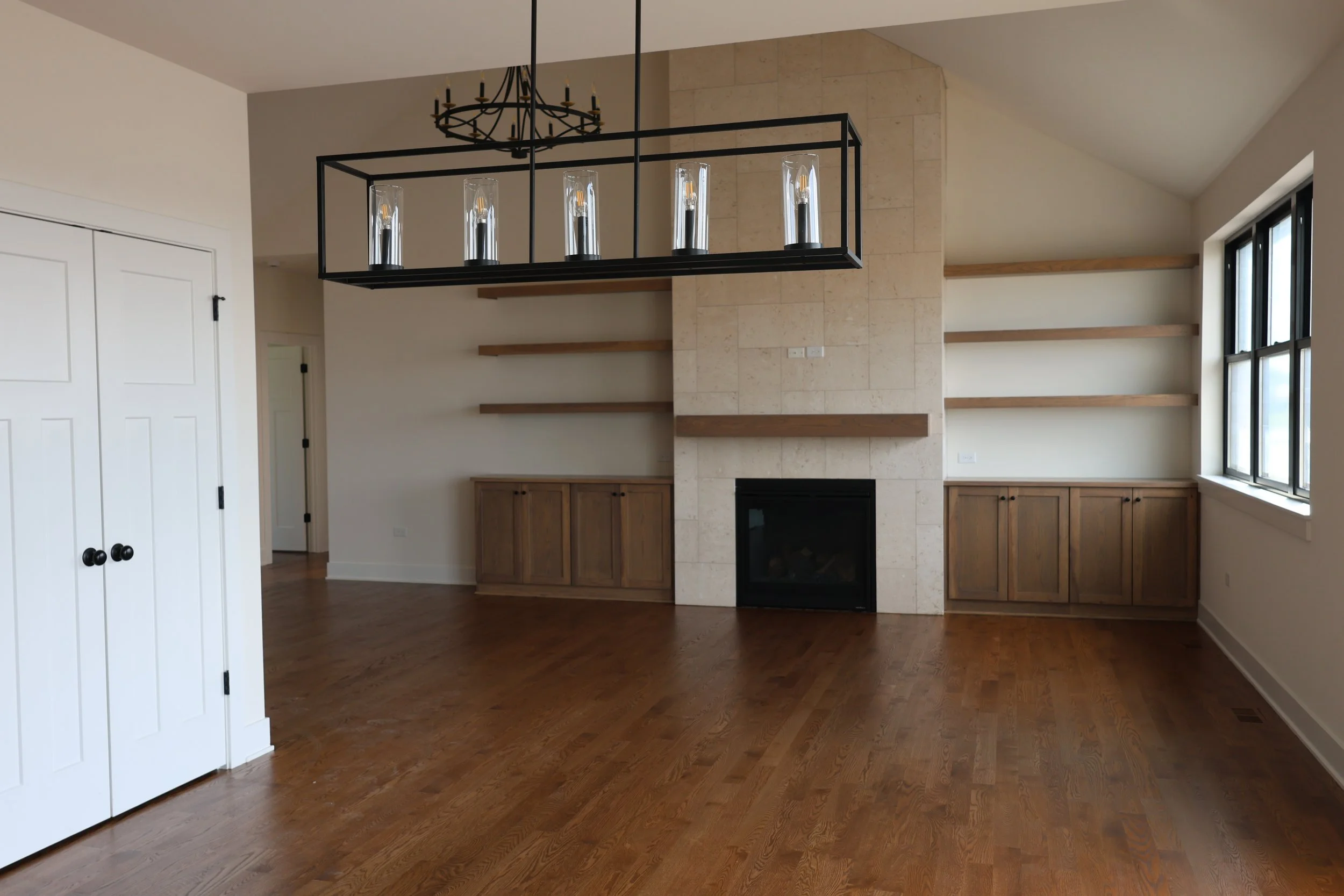Empty living room with hardwood floors, built-in wooden cabinets, a fireplace, open shelves, large window, and a modern black chandelier.