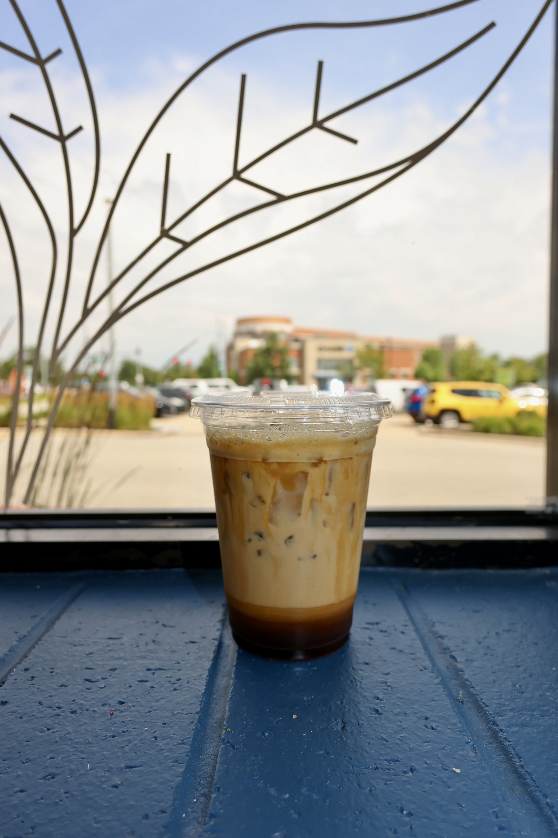 A plastic cup of iced caramel coffee on a dark blue table by a window with a view of parking lot and a building in the background.