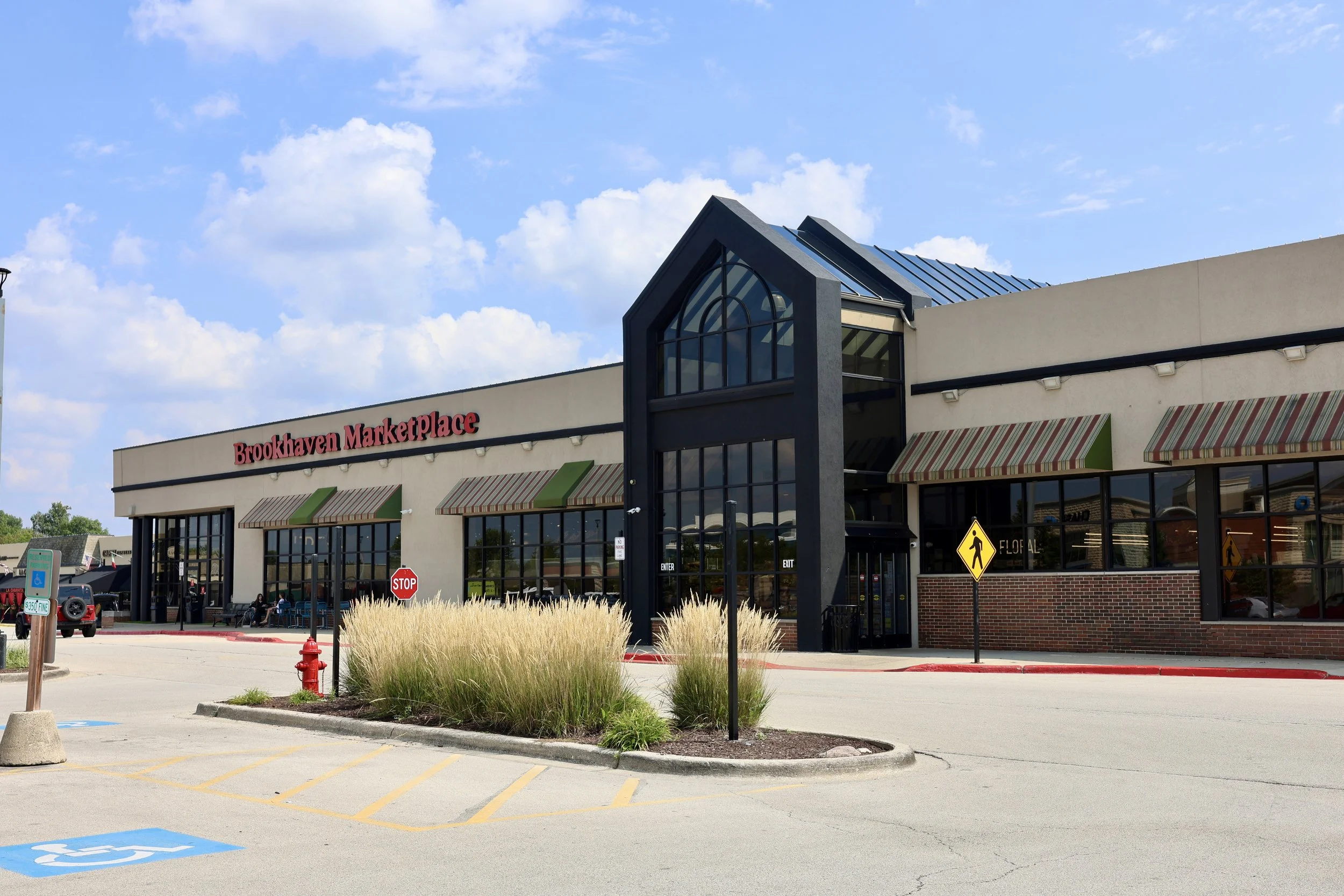 Exterior of Brookhaven Marketplace shopping center with parking lot, striped awnings, and a blue roofed glass entrance under a partly cloudy sky.