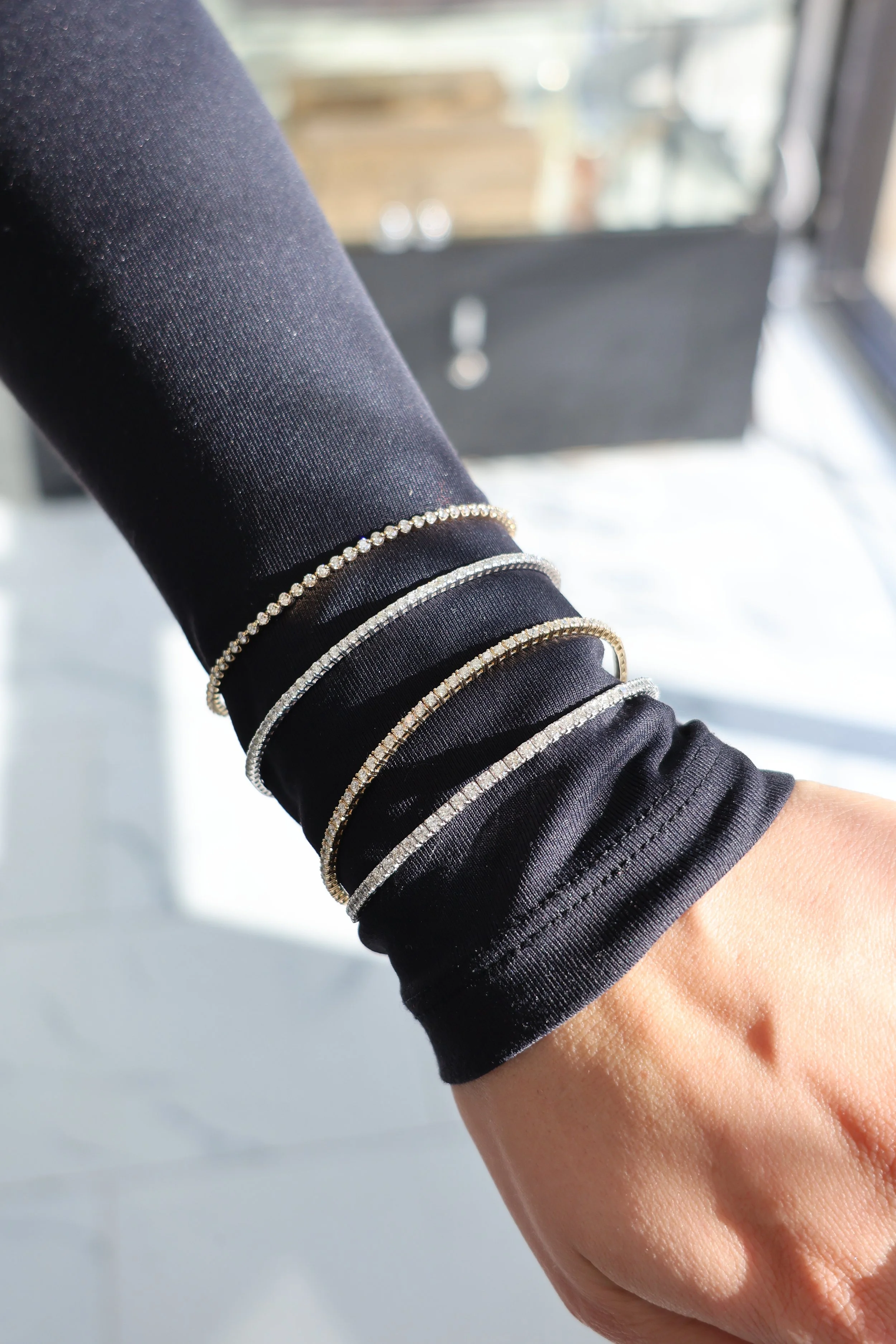 Close-up of a person's arm with multiple diamond bracelets, wearing a black long-sleeve shirt, near a window with sunlight.
