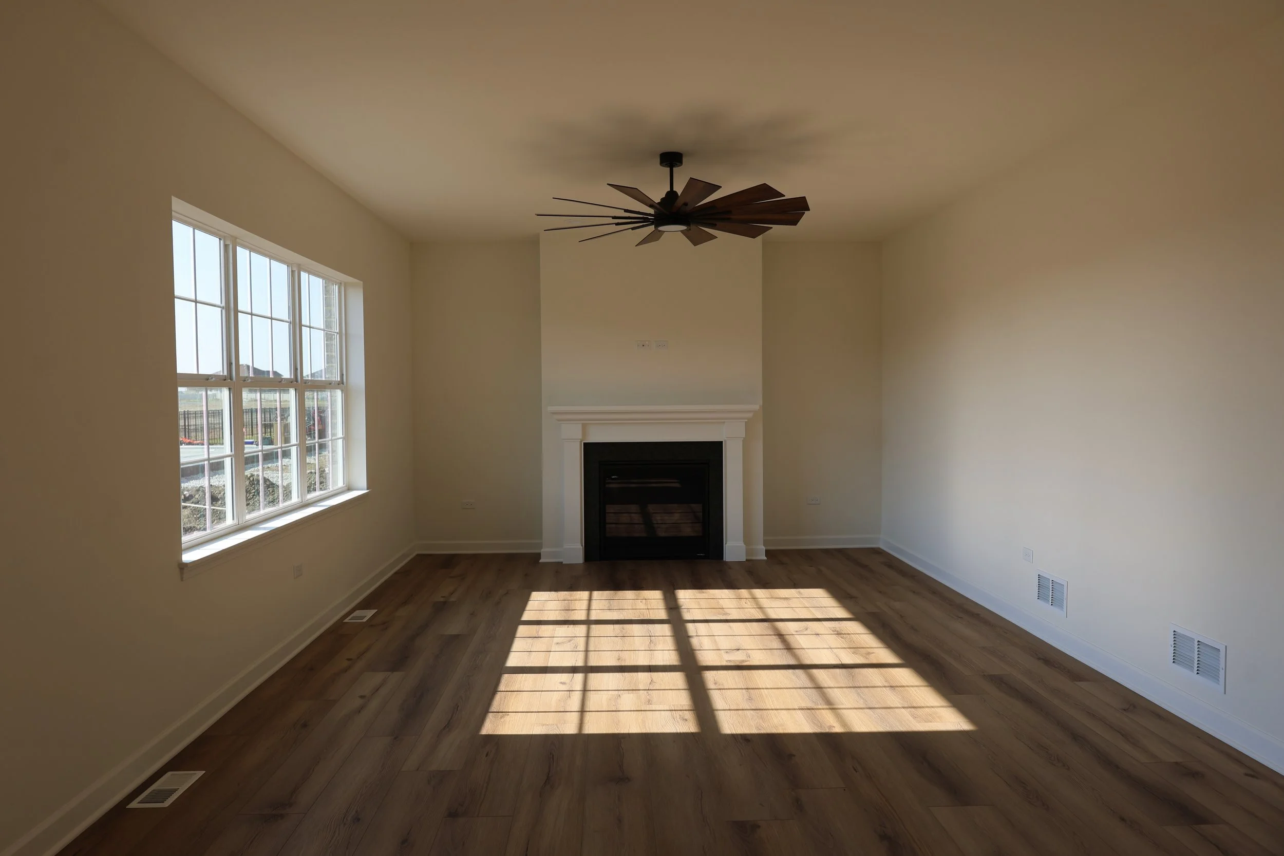 Empty living room with large front window, wooden floor, a white fireplace on the wall, ceiling fan, and sunlight casting shadows through the window.